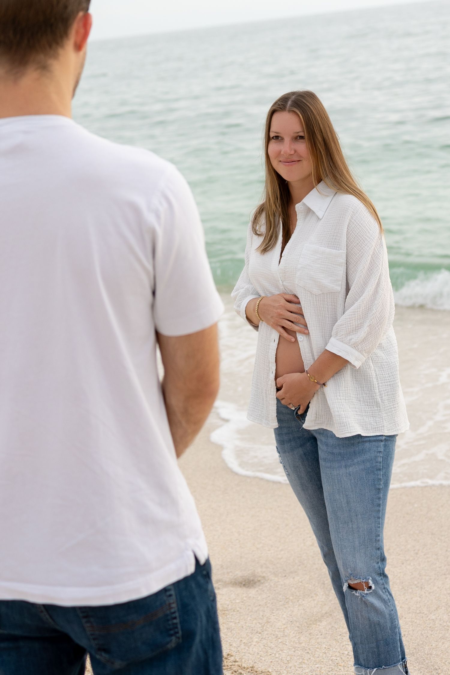Pregnant woman in white shirt cradling baby bump on beach while partner stands in foreground by ocean.
