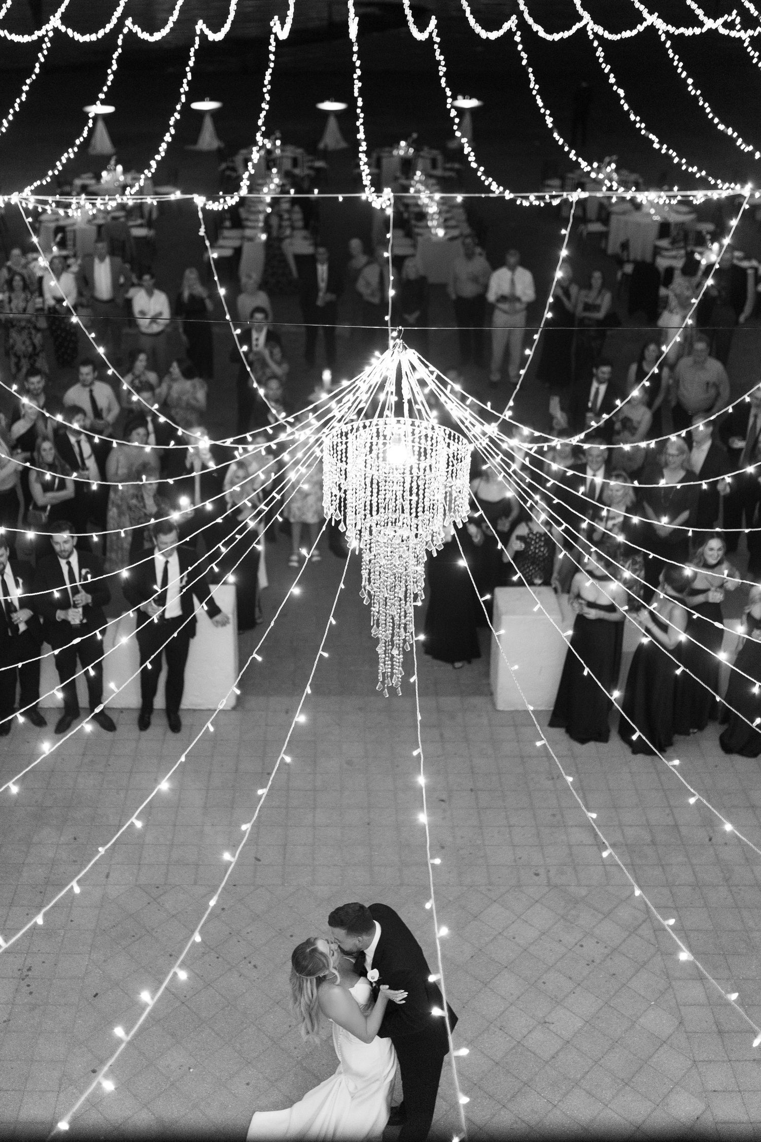 Black and white aerial view of bride and groom sharing first dance under sparkling chandelier and lights.