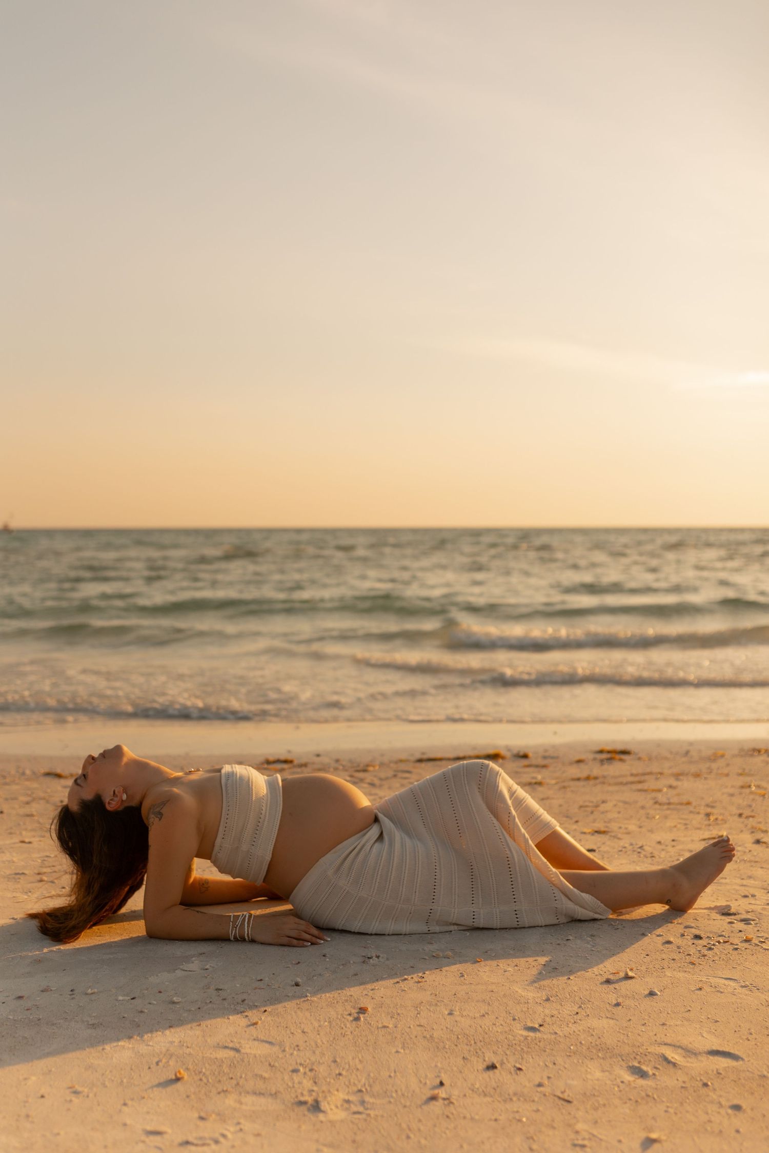 Pregnant woman in white outfit lies on sandy beach at sunset with ocean waves in background.
