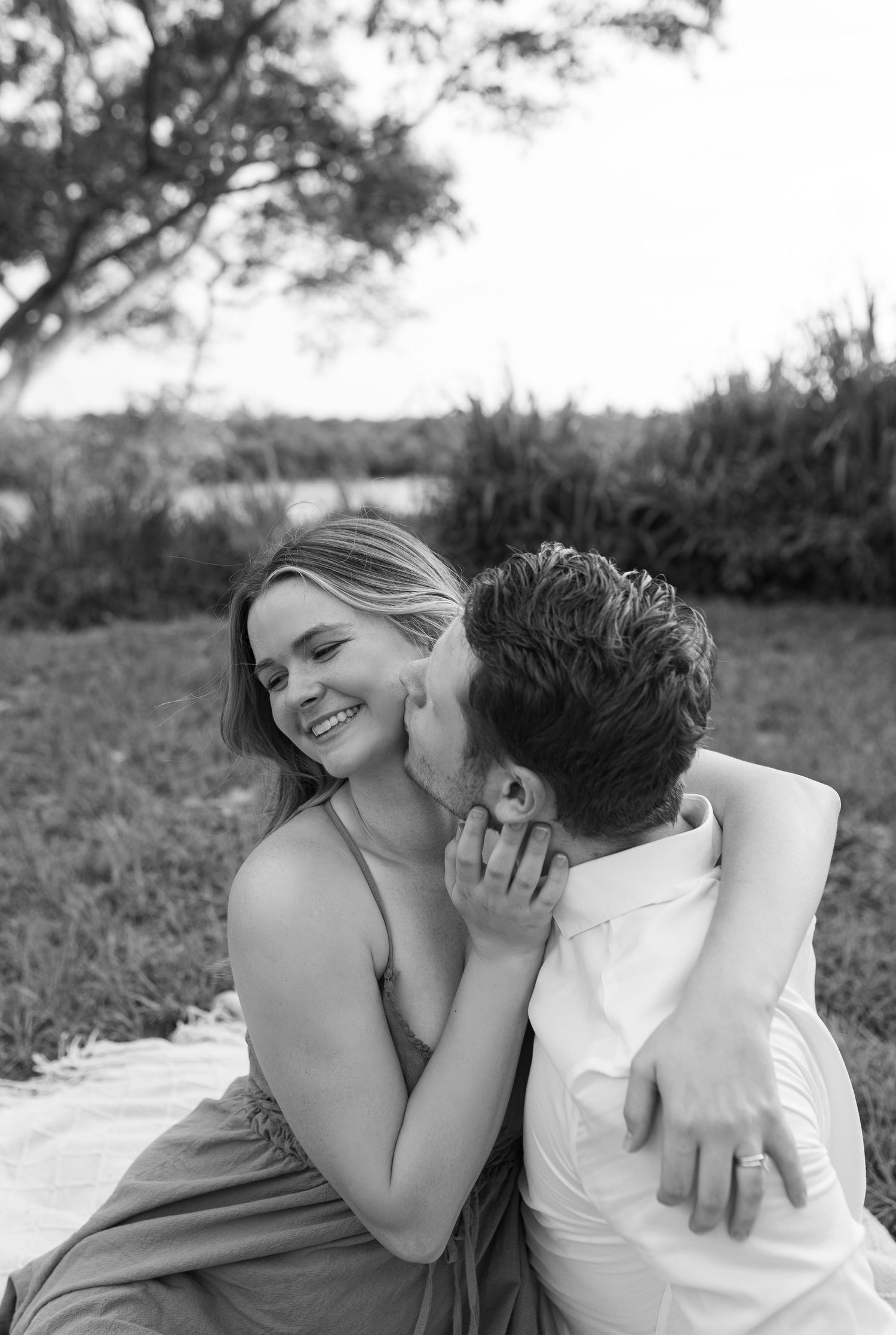 Black and white photo of smiling woman being kissed on cheek by man while sitting outdoors.