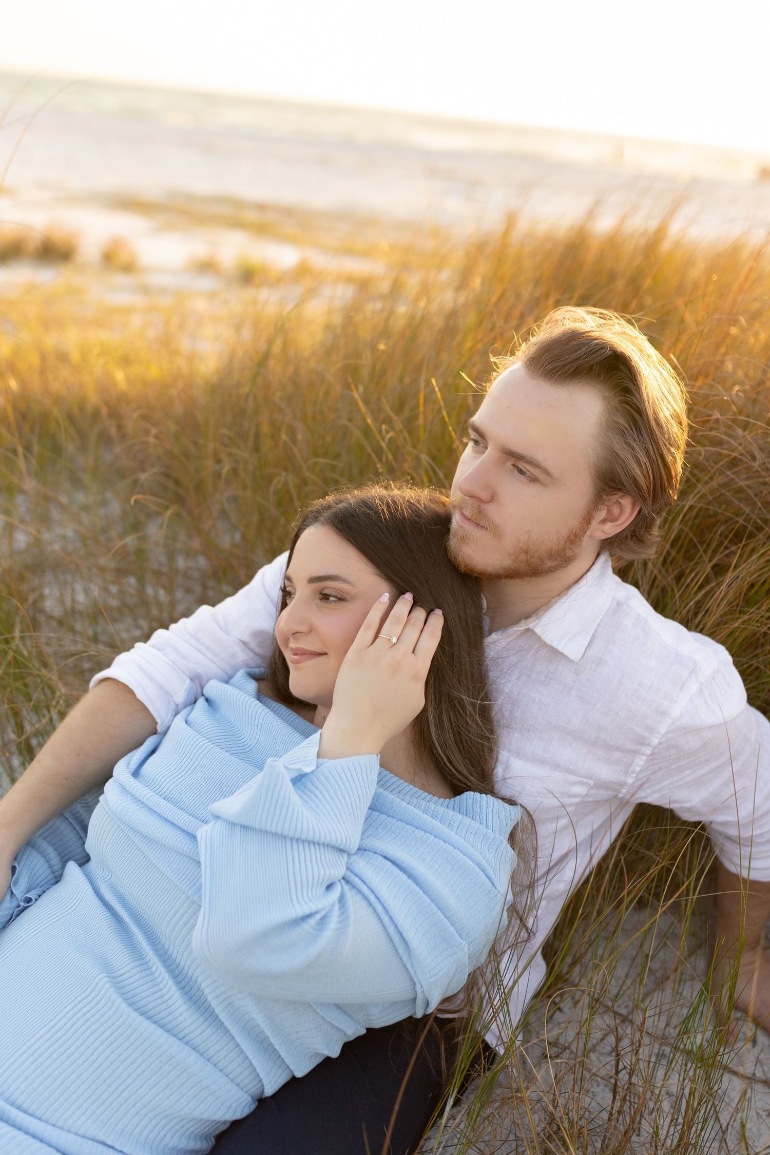 Couple relaxing in golden hour sunlight among beach dunes, woman resting her head on mans shoulder tenderly.
