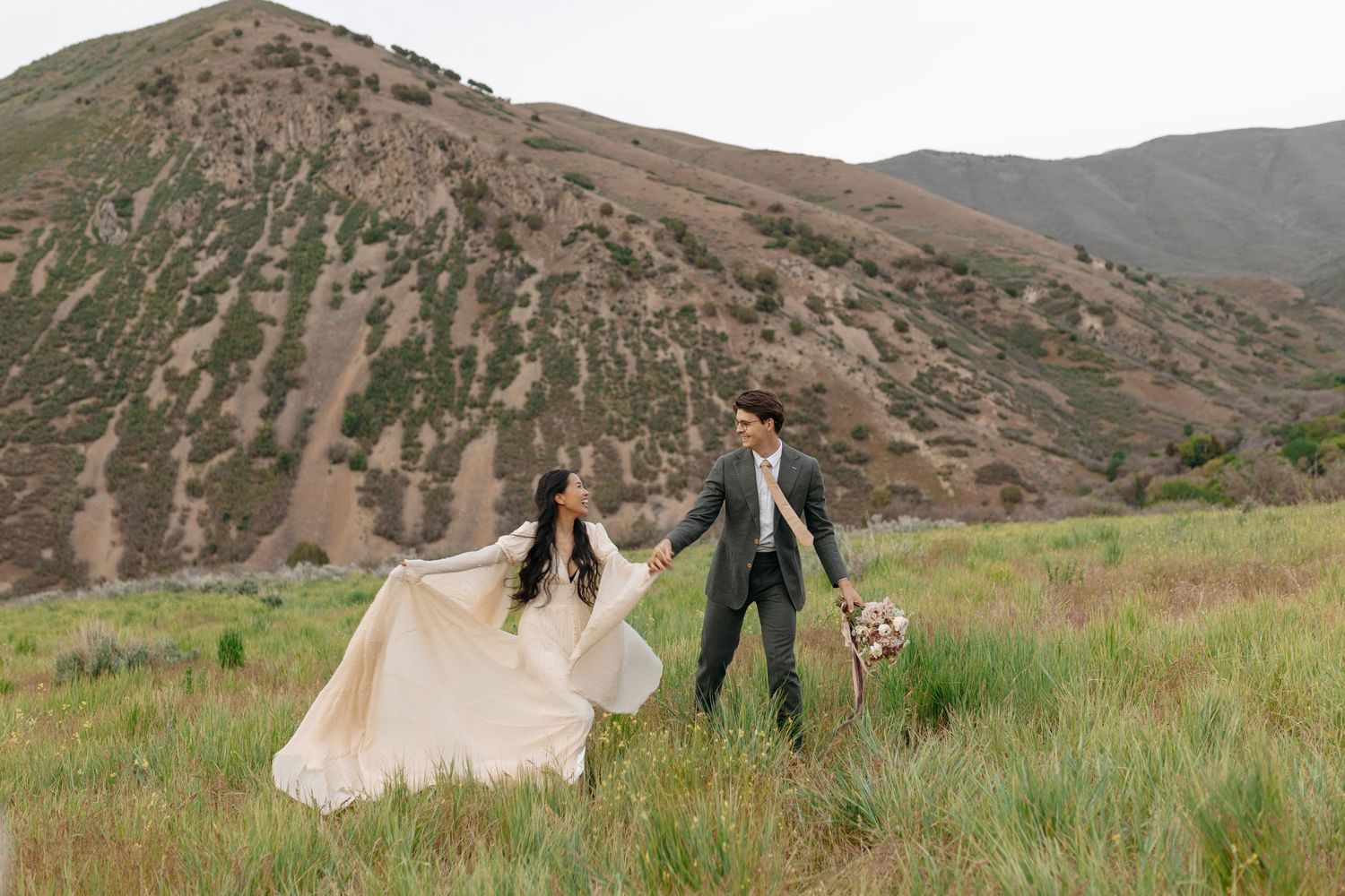 Bride and groom holding hands walking through green mountain meadow, brides gown flowing in the wind.