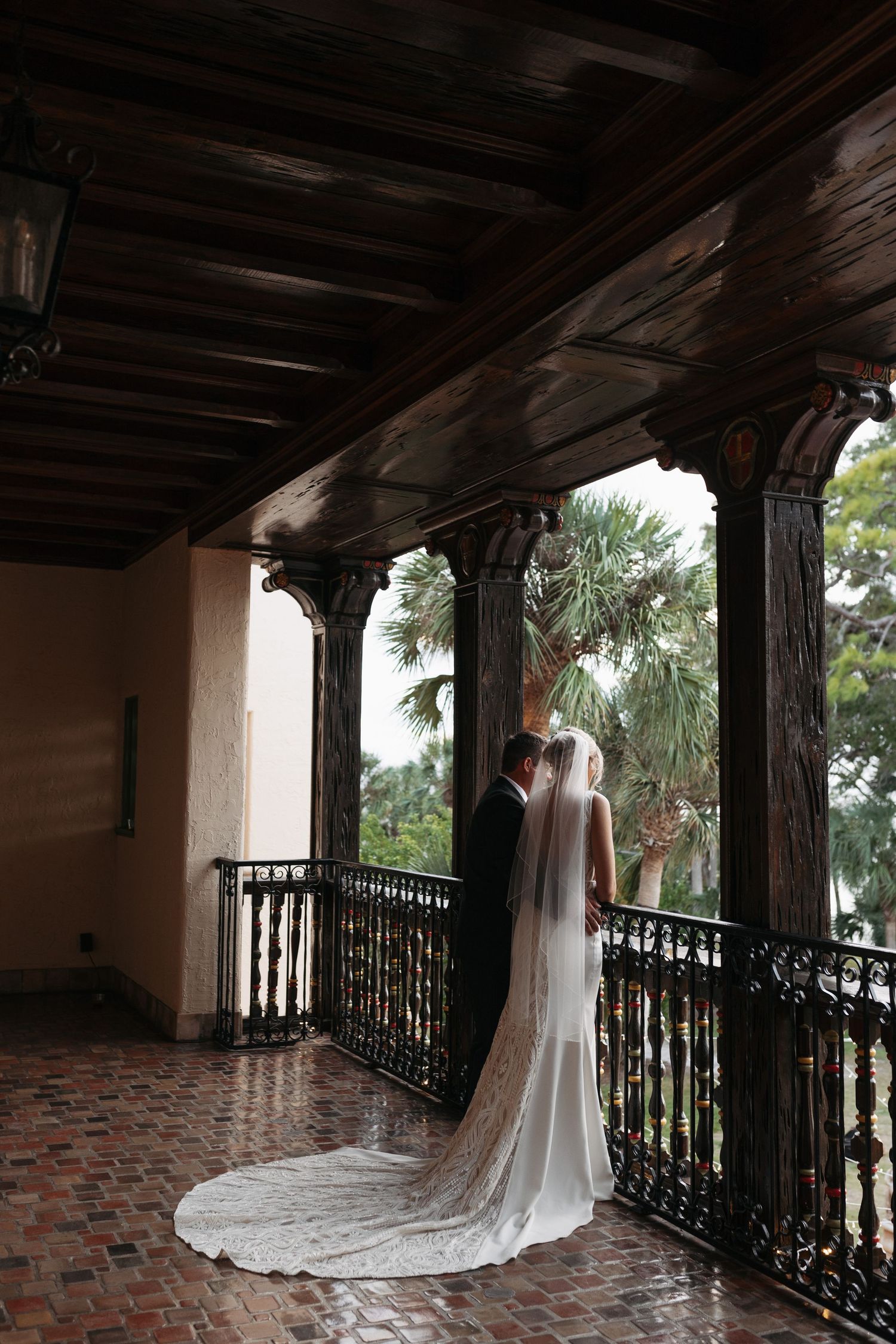 Bride and groom stand on ornate dark wood balcony with iron railings overlooking tropical greenery.