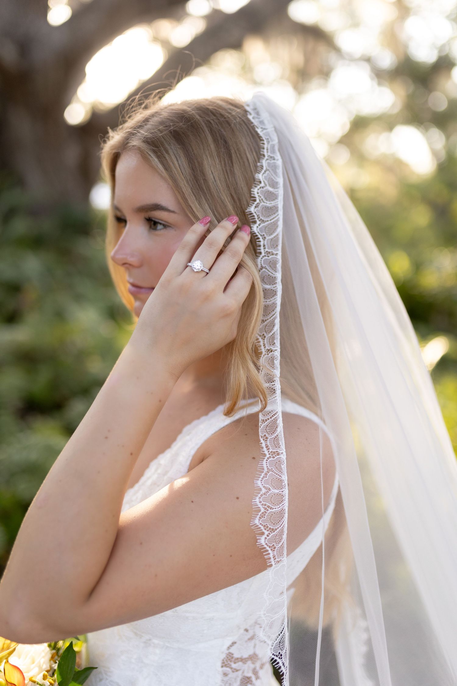 Blonde bride in white lace gown wearing veil and diamond ring touching hair in golden light.