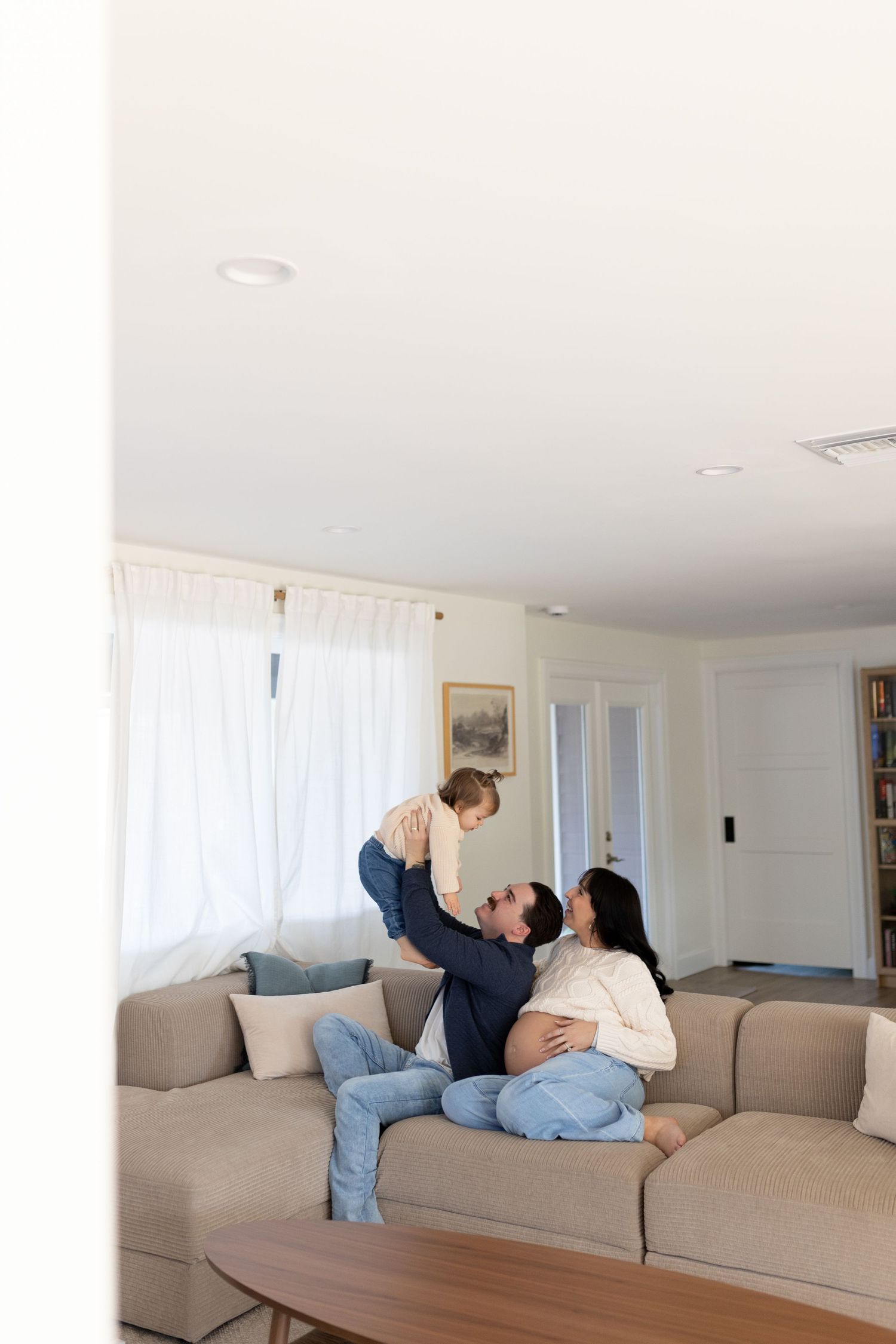 Family relaxing on a couch as father lifts toddler while pregnant mother reclines nearby.