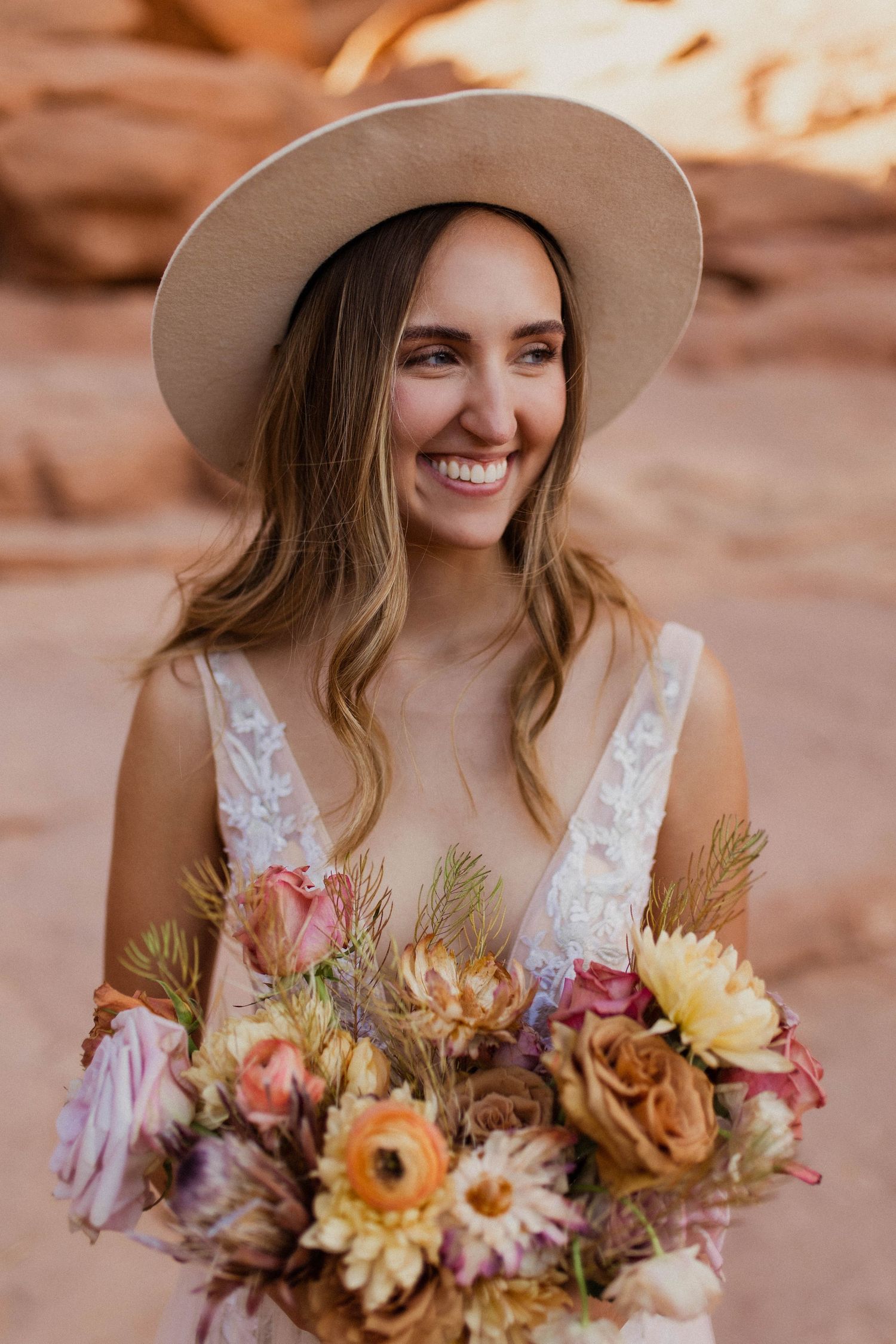 Joyful boho bride in wide brim hat holding a vibrant mixed floral bouquet against red rock formations.