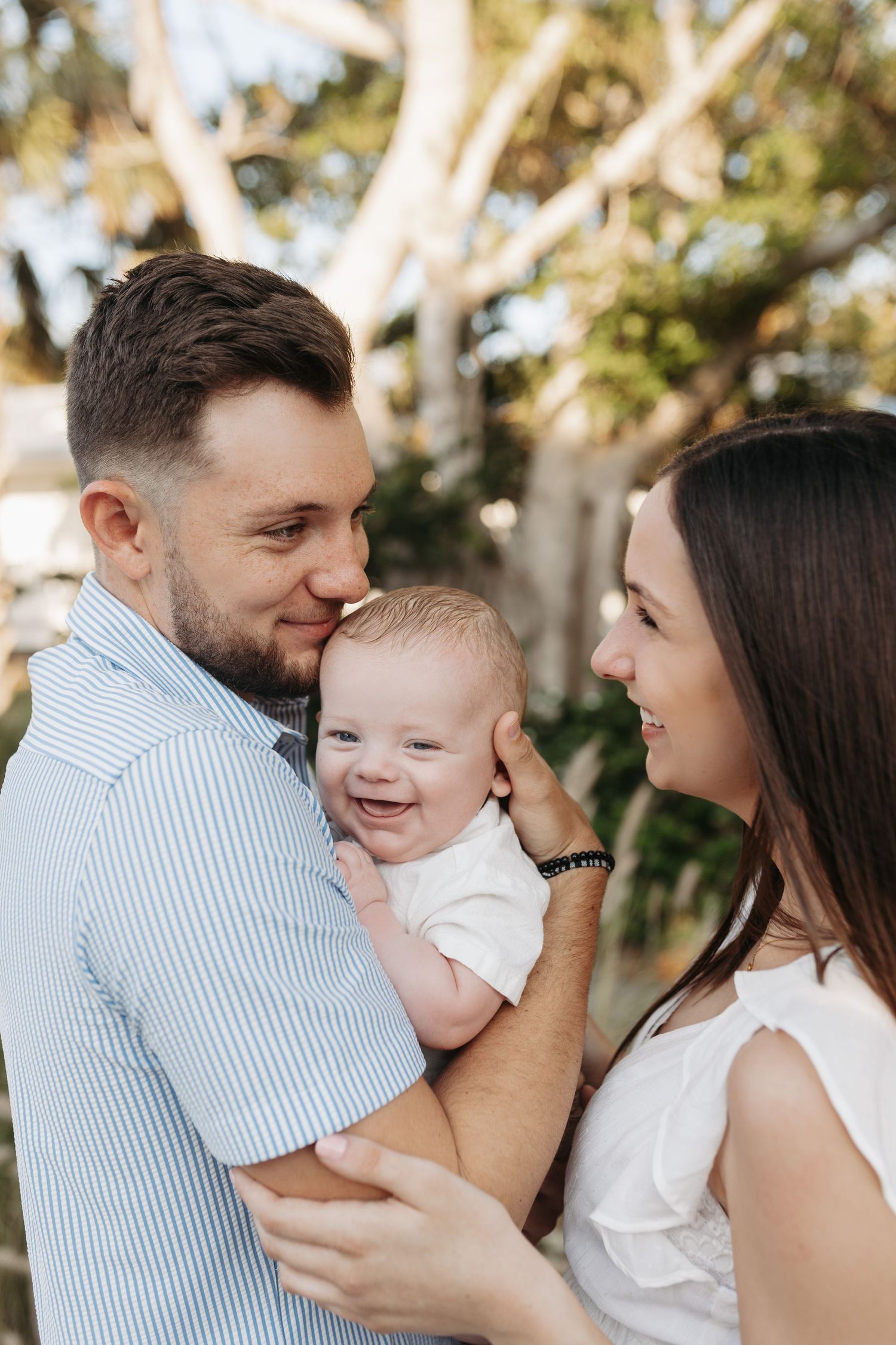 Smiling parents holding their laughing baby outdoors surrounded by lush green trees.