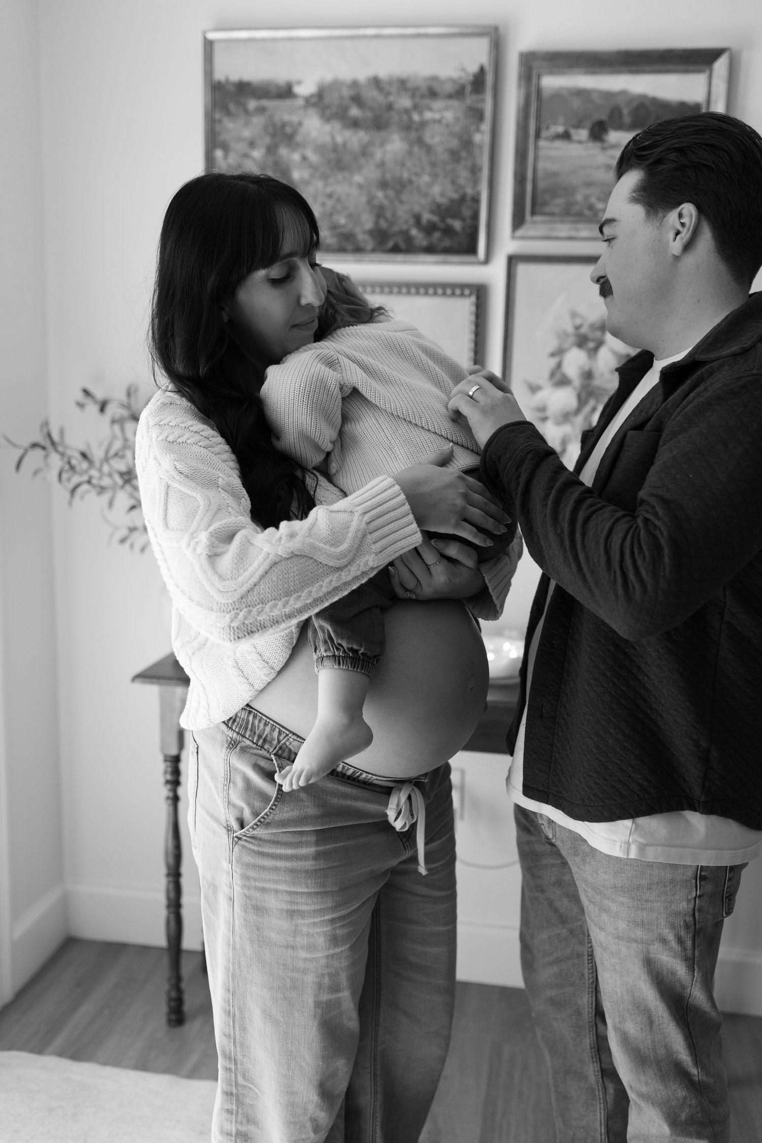 Black and white maternity photo of a couple tenderly holding a pregnant belly indoors.