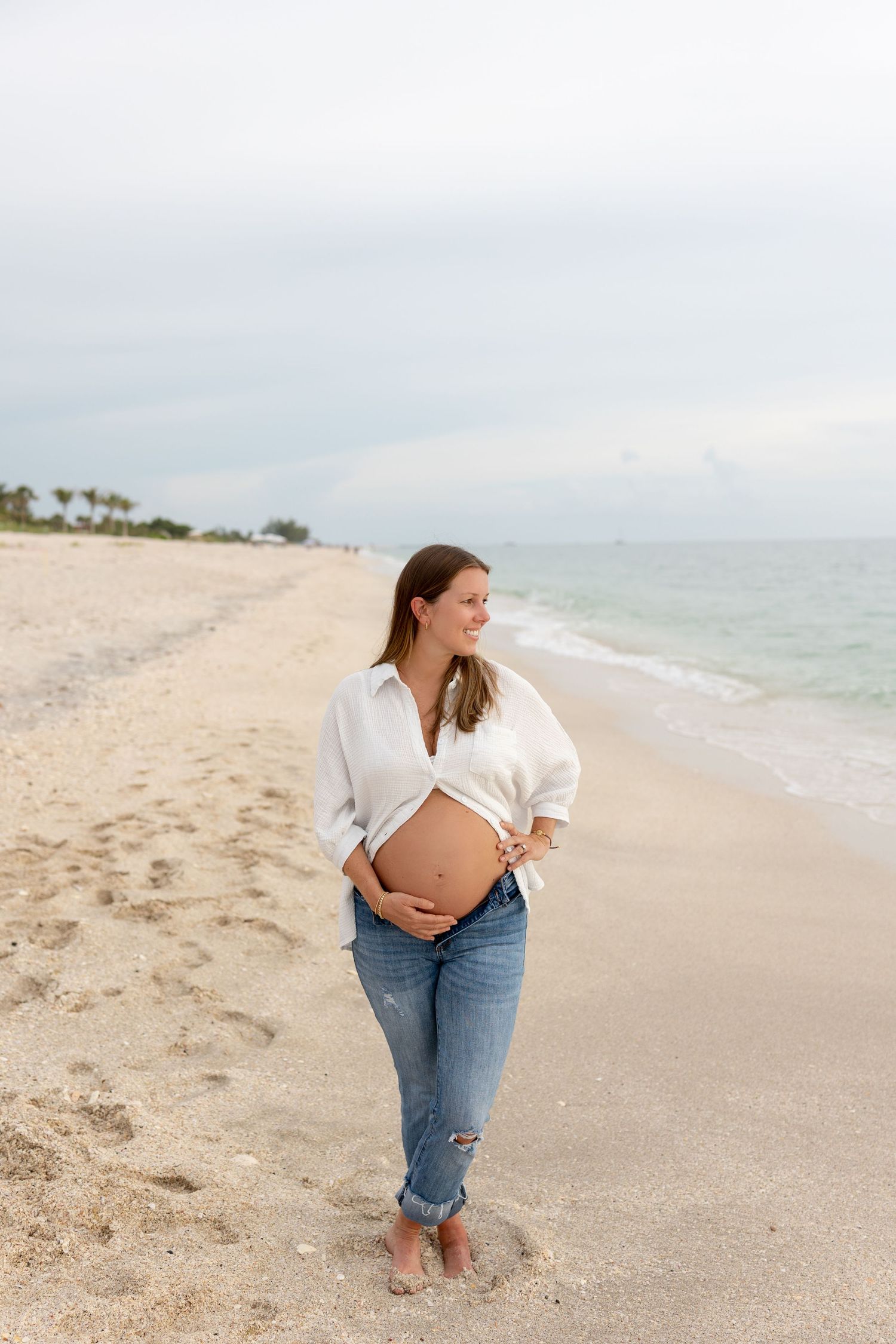 Pregnant woman in white shirt and jeans cradles her baby bump while standing on a sandy beach.
