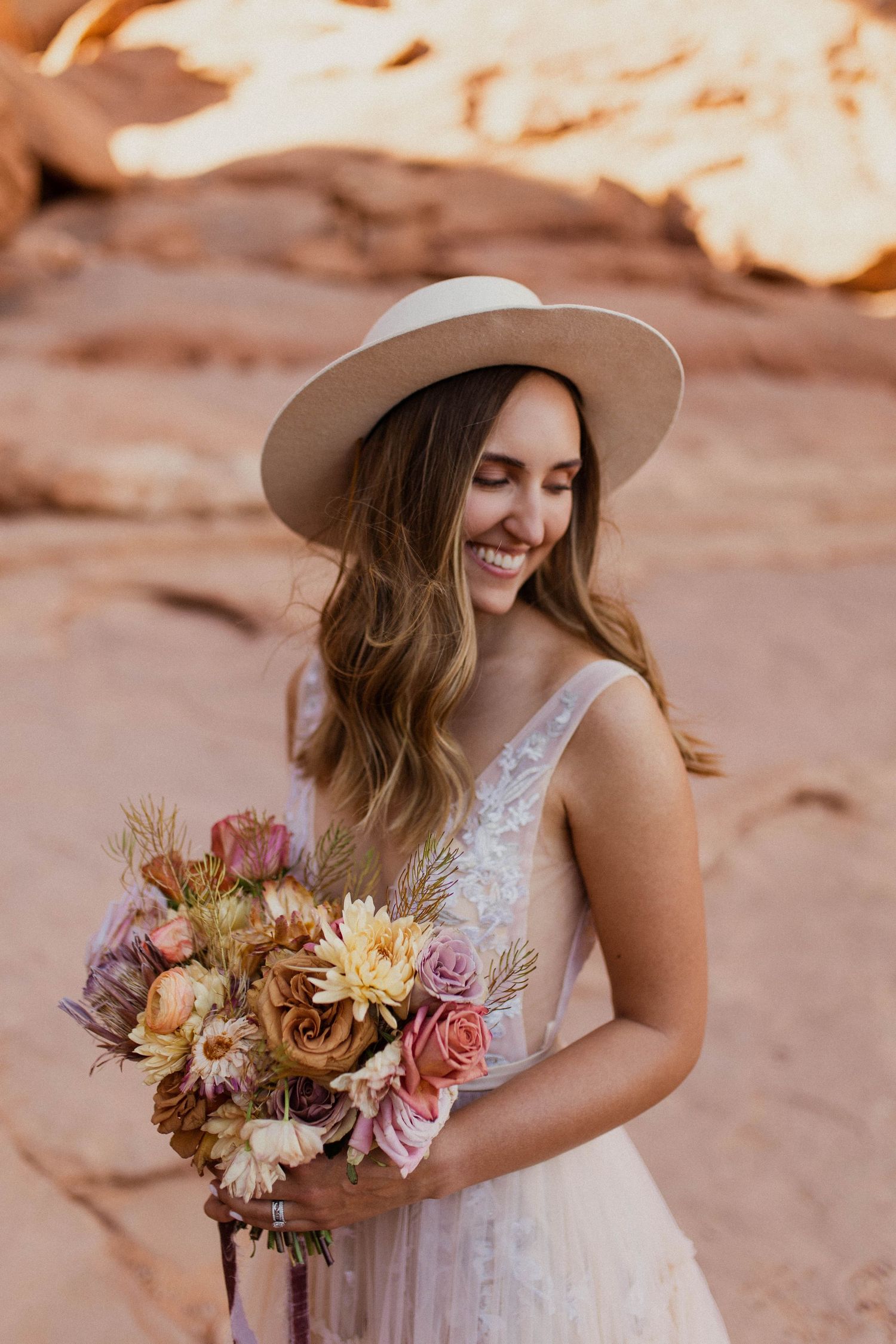 Smiling boho bride in white lace dress and wide brim hat holding colorful desert wildflower bouquet.