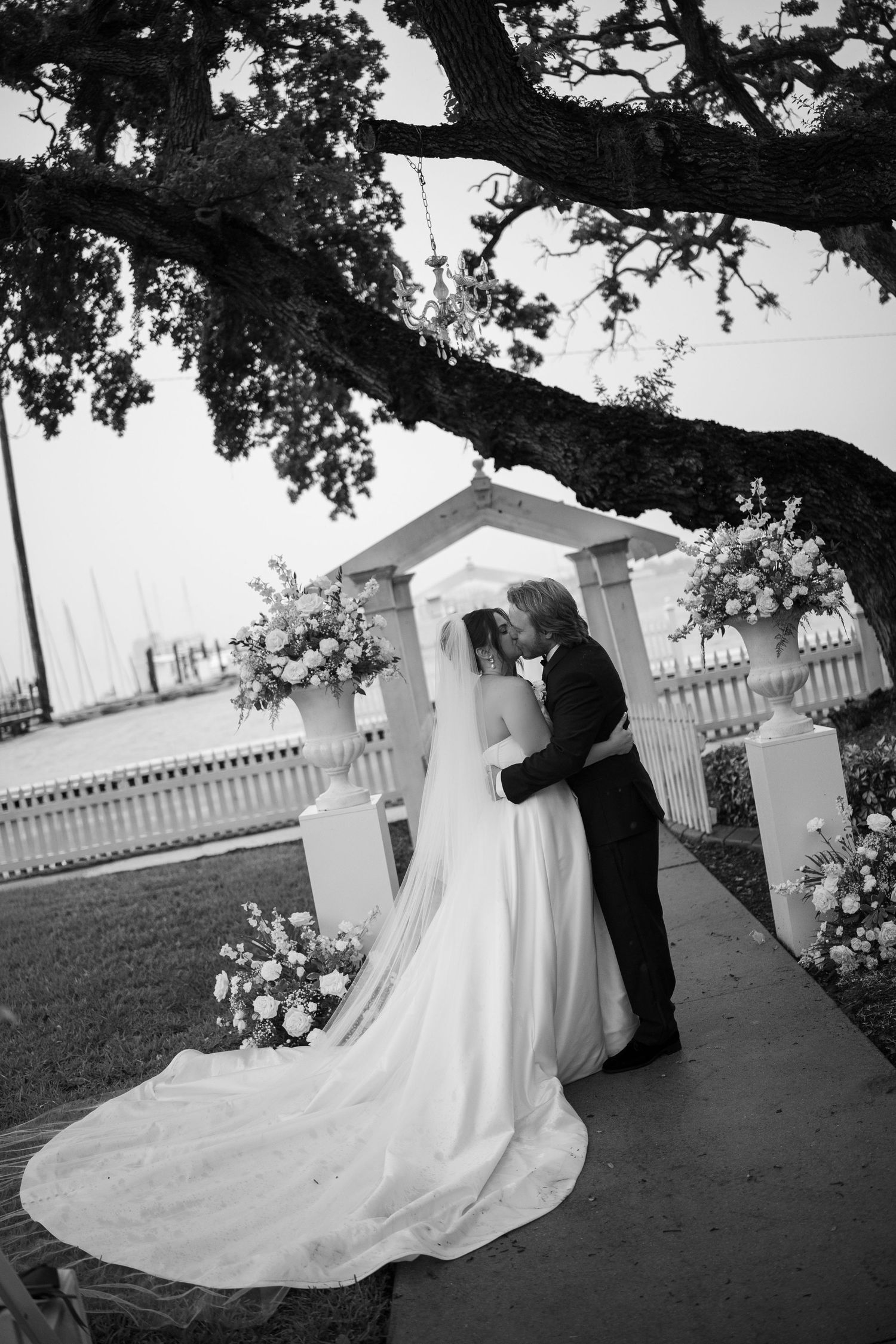 Black and white wedding photo of bride and groom kissing under oak tree at outdoor altar with floral arrangements.