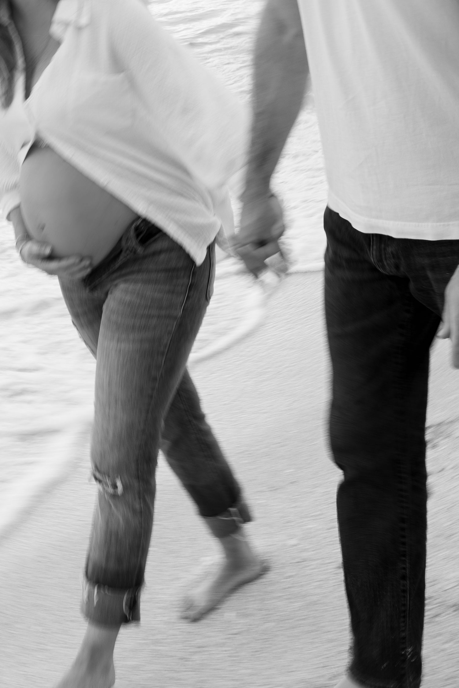 Black and white motion blur photo of pregnant couple holding hands walking on beach.