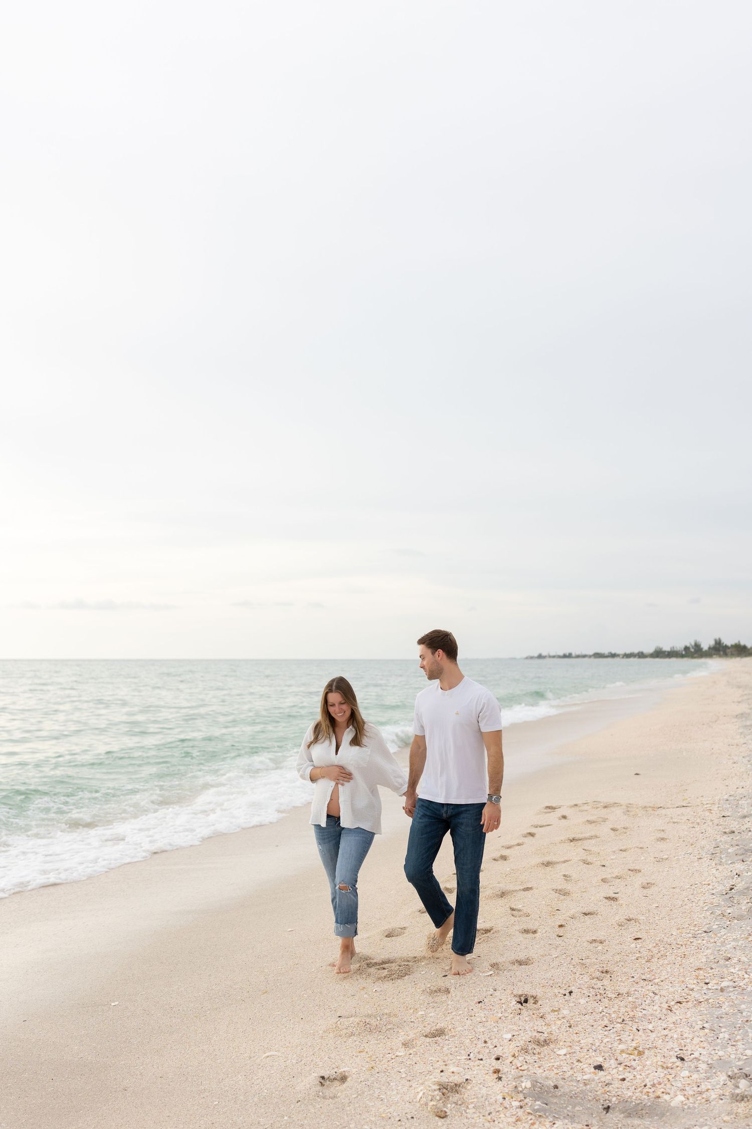 Couple in white shirts and jeans walking hand in hand along sandy beach with calm ocean waves.
