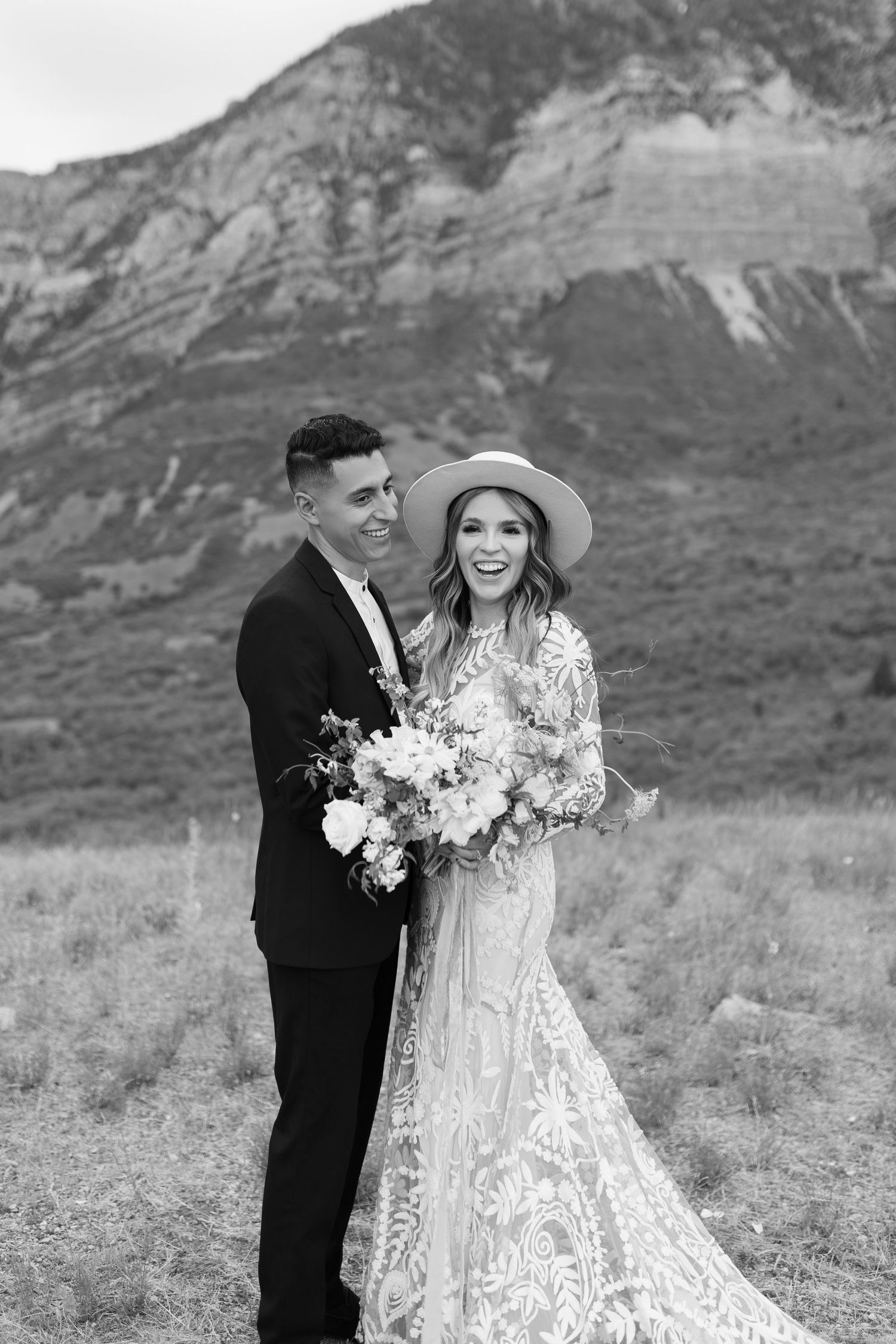 Black and white portrait of smiling newlyweds with mountain backdrop, bride holding a lush floral bouquet.