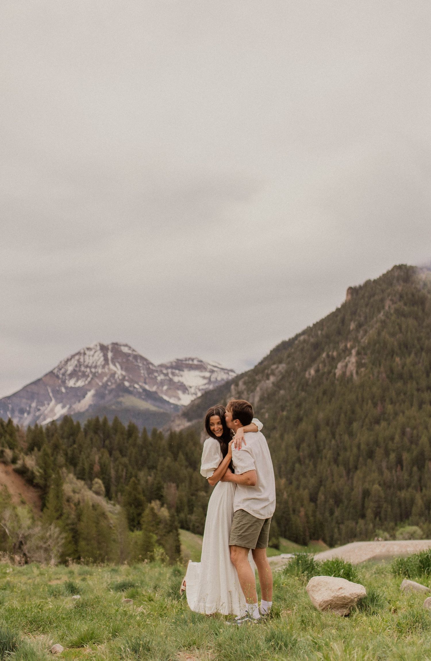 Couple embracing in a lush green mountain meadow with snow-capped peaks and pine forest behind them.