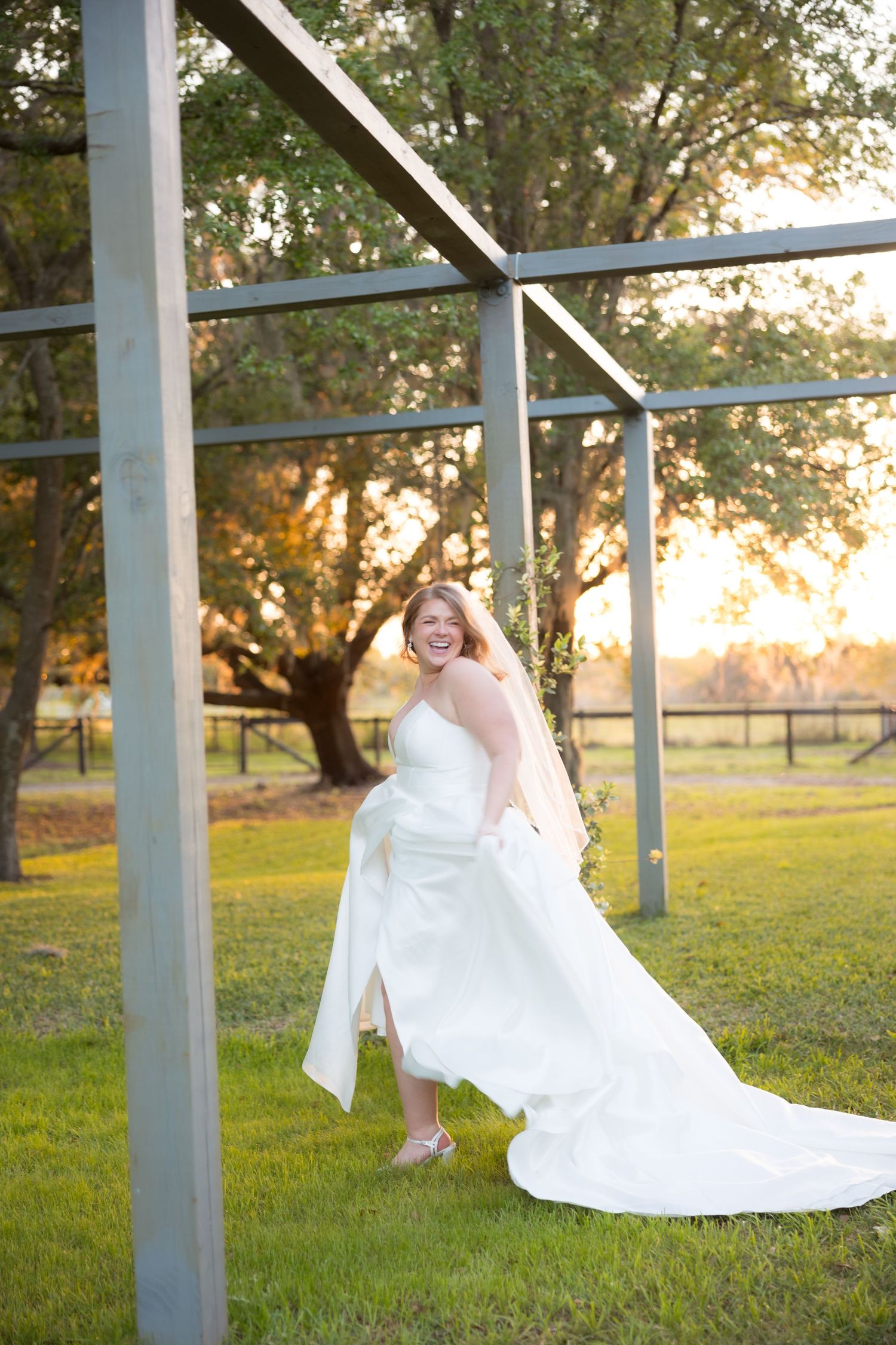 Bride in a white gown laughing and twirling near a metal pergola at golden hour sunset.