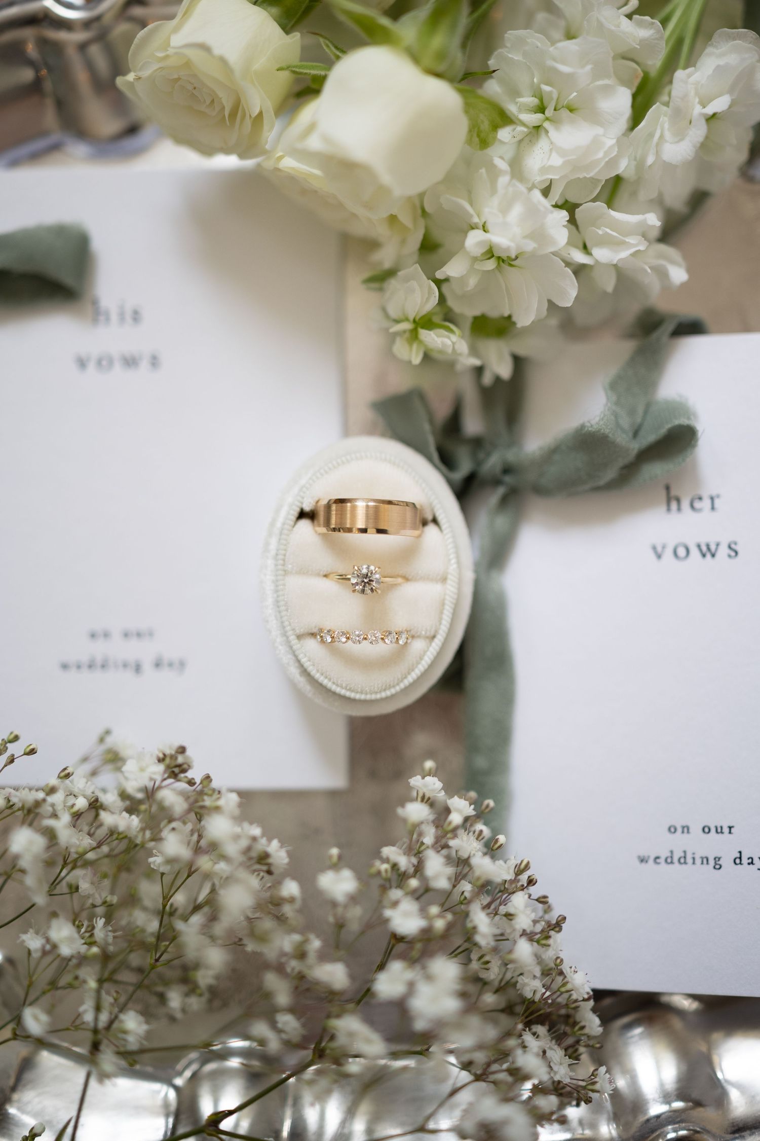 Wedding flat lay featuring gold rings in velvet box surrounded by white roses, gypsophila, and vow booklets.
