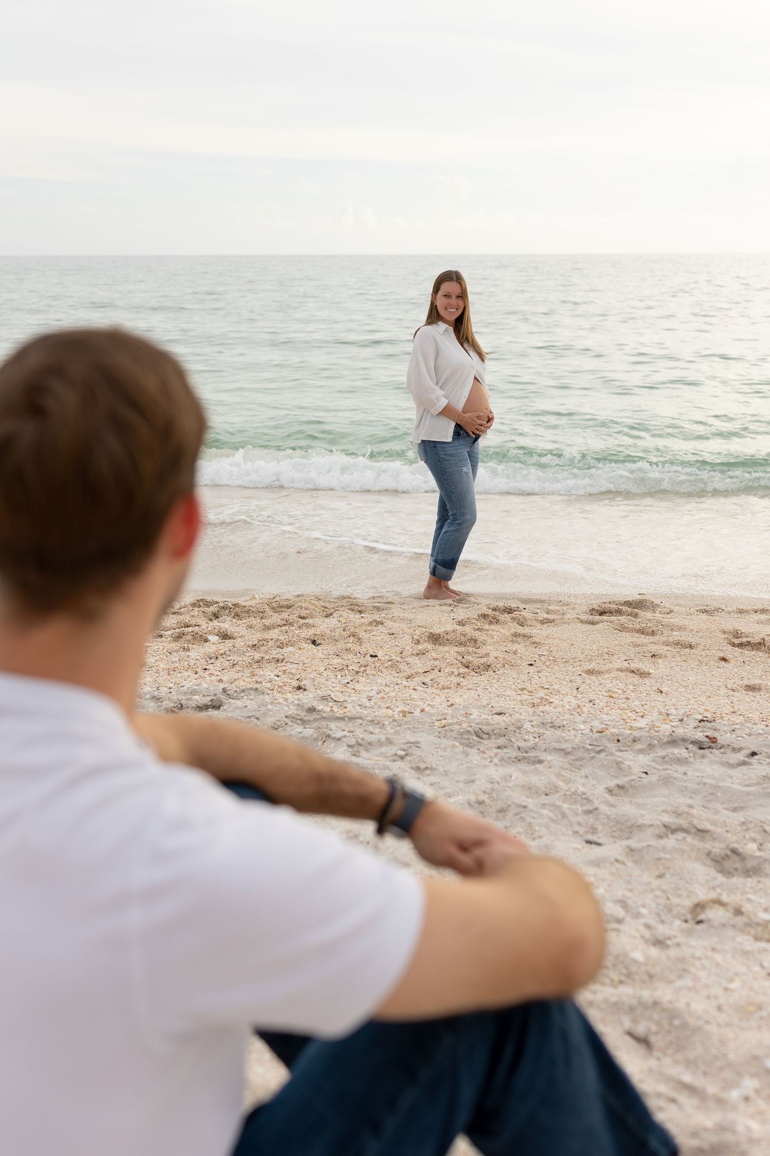 Pregnant woman in white shirt and jeans stands at ocean's edge while partner watches from beach.