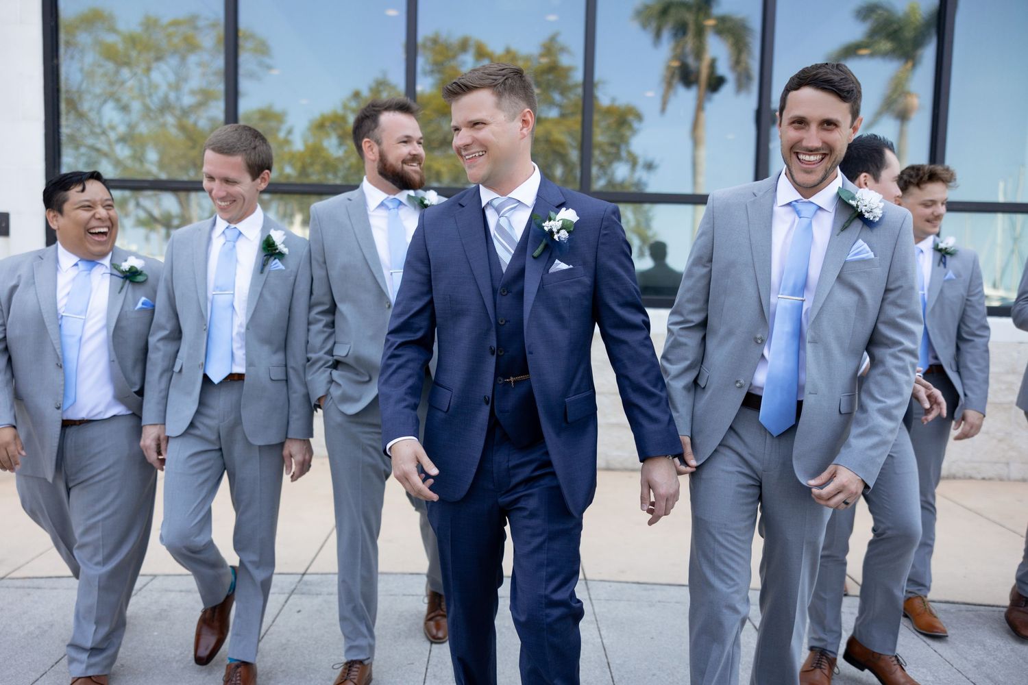 Groom in navy three-piece suit walks confidently with smiling groomsmen in grey suits outside venue.