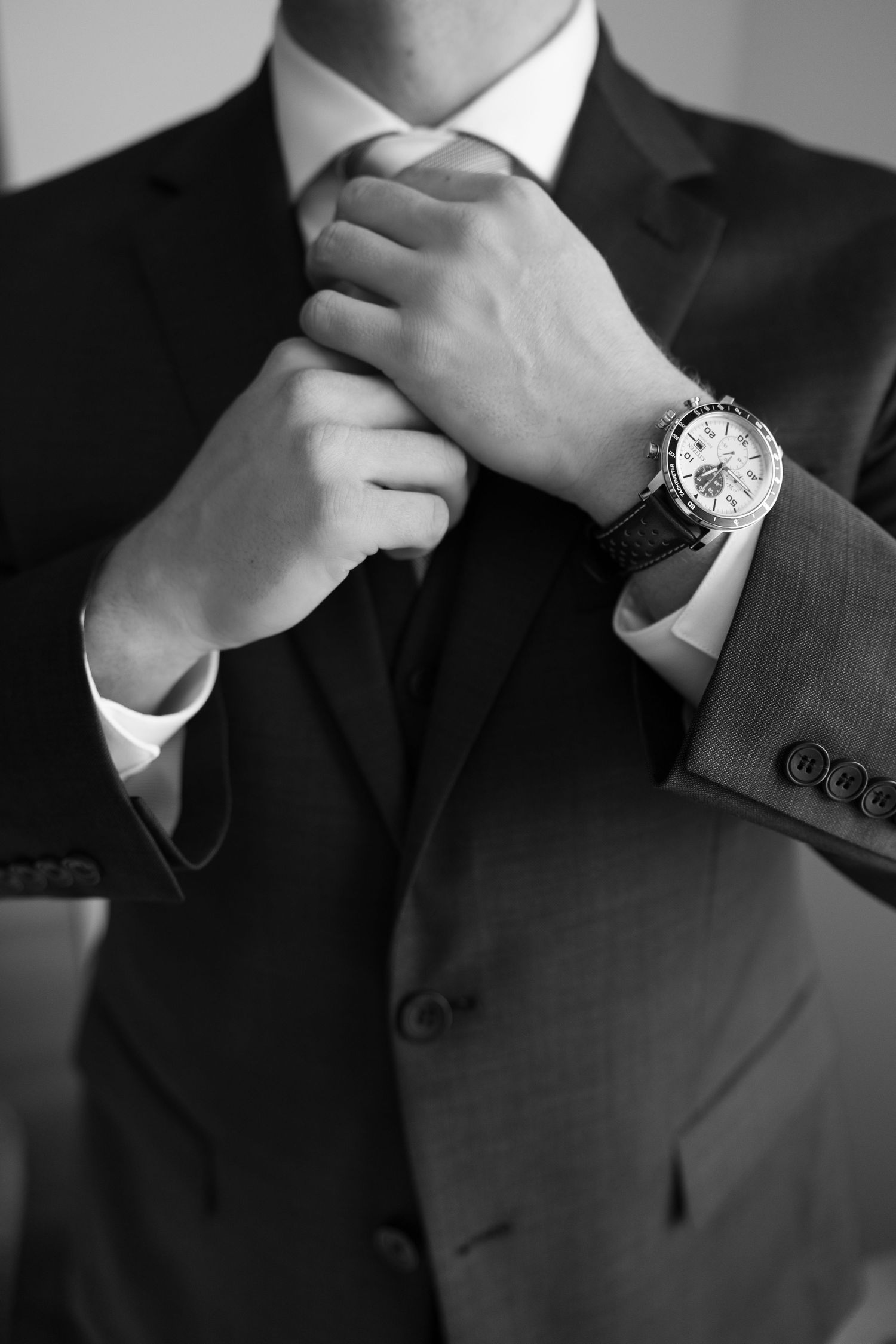 Black and white close-up of a groom adjusting his tie while wearing a luxury watch on his wedding day.