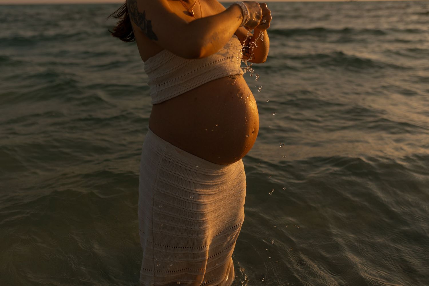 A pregnant woman in a sheer white wrap stands in ocean water at golden hour sunset.