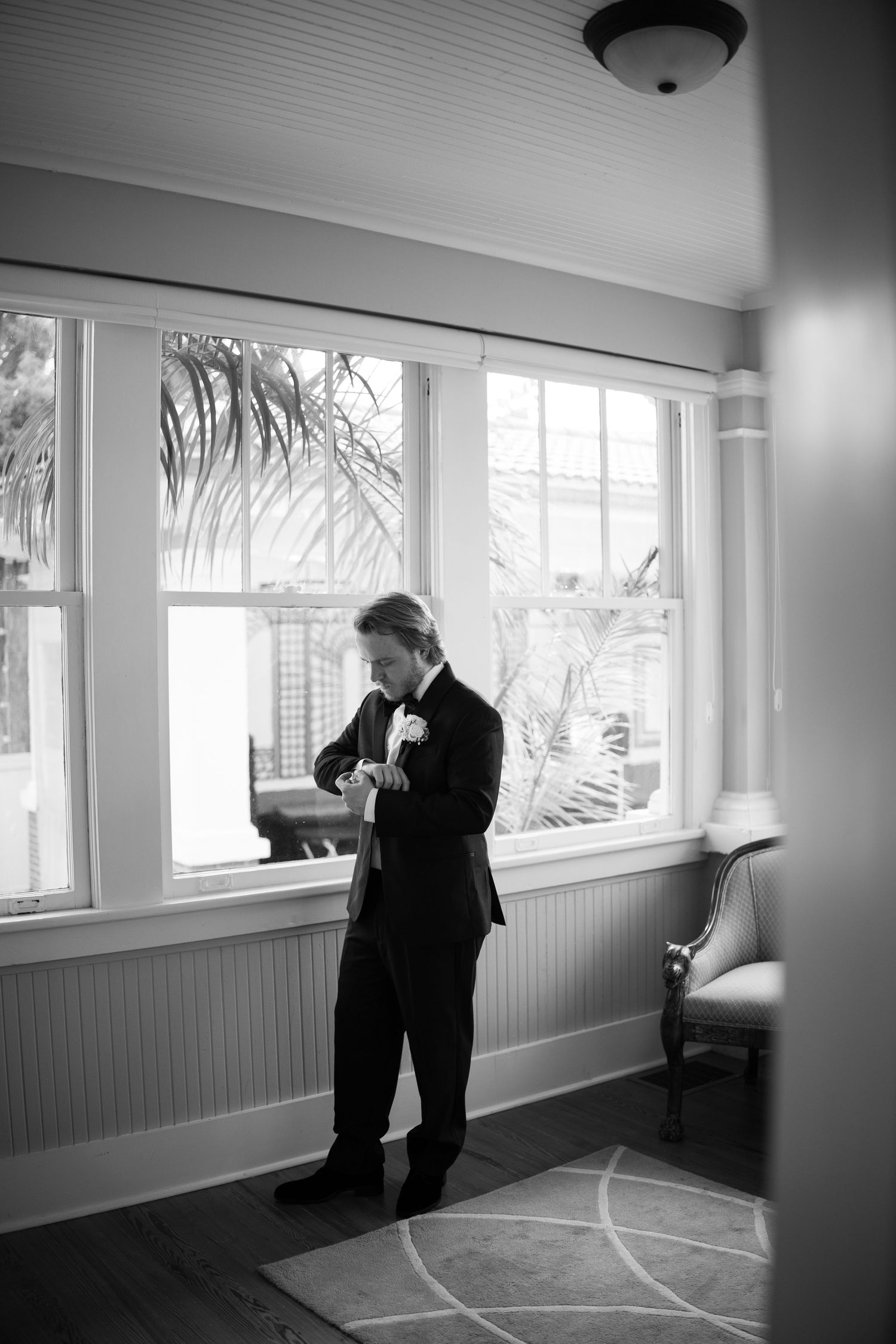 Black and white photo of groom in tuxedo checking watch by large windows in bright sunlit room.