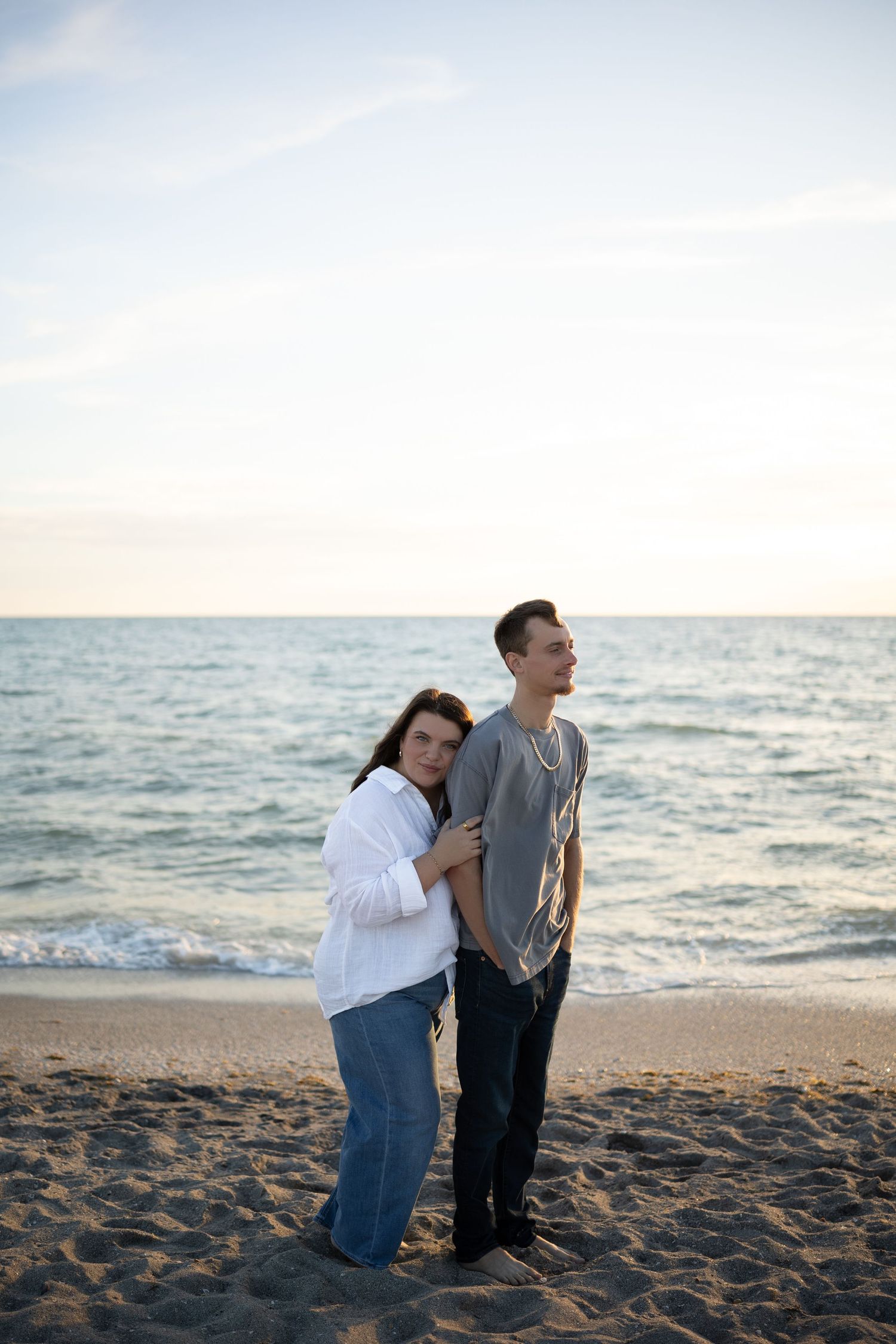 Couple stands embracing on a sandy beach with calm ocean waves at sunset behind them.
