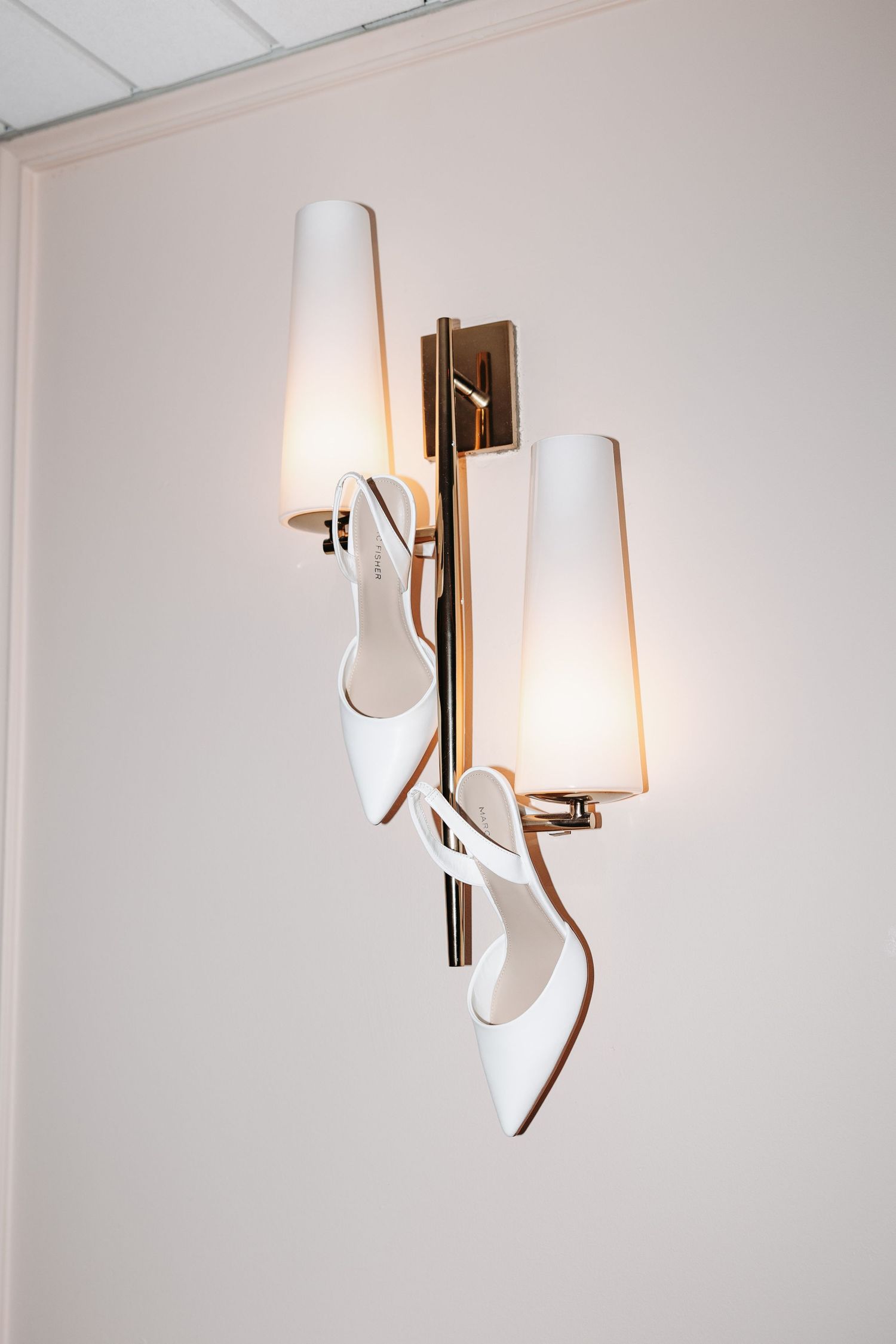 White bridal heels hang decoratively on a gold wall sconce with frosted glass shades.