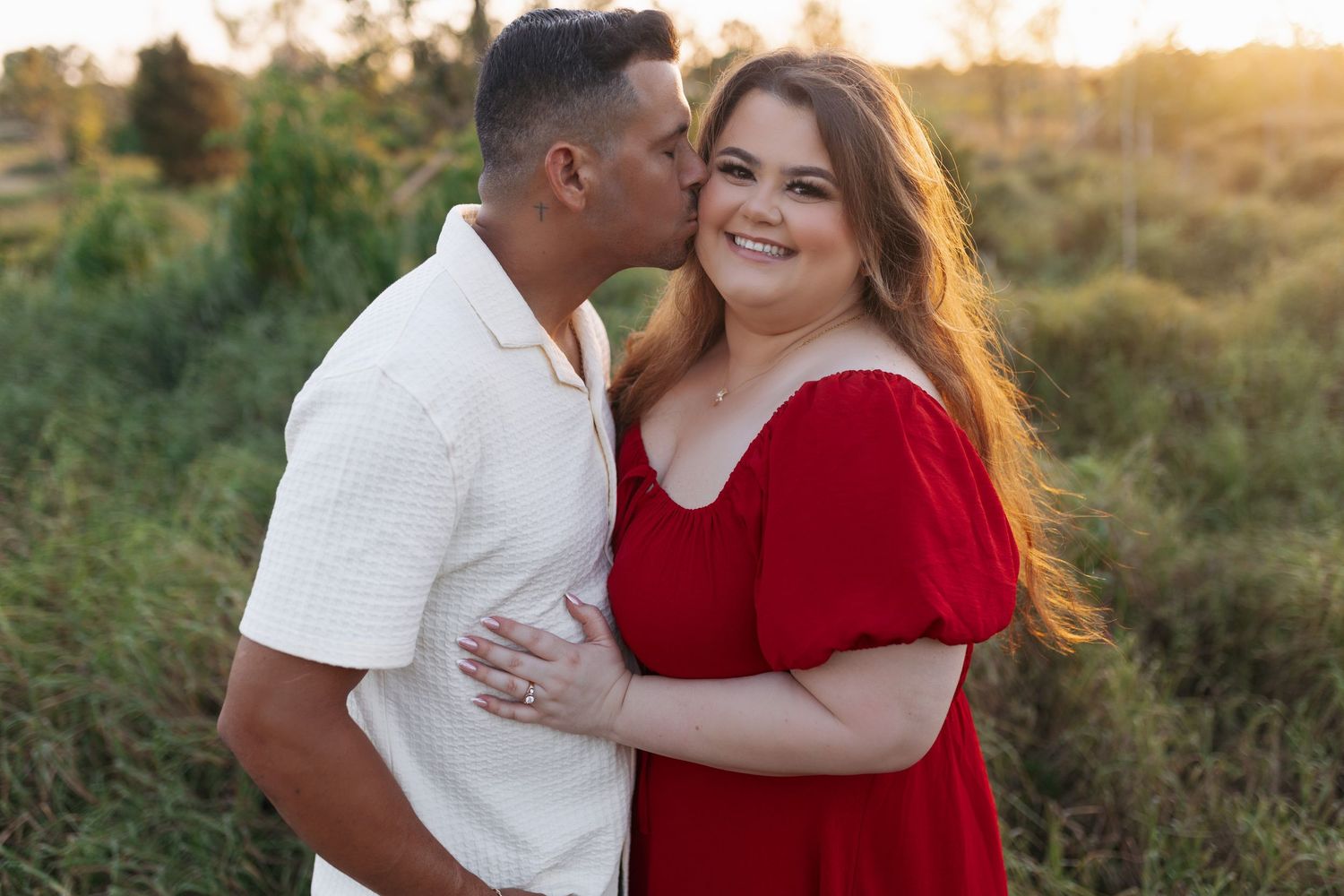 Man in white shirt kissing woman in red dress on the cheek during a sunset engagement photo session.