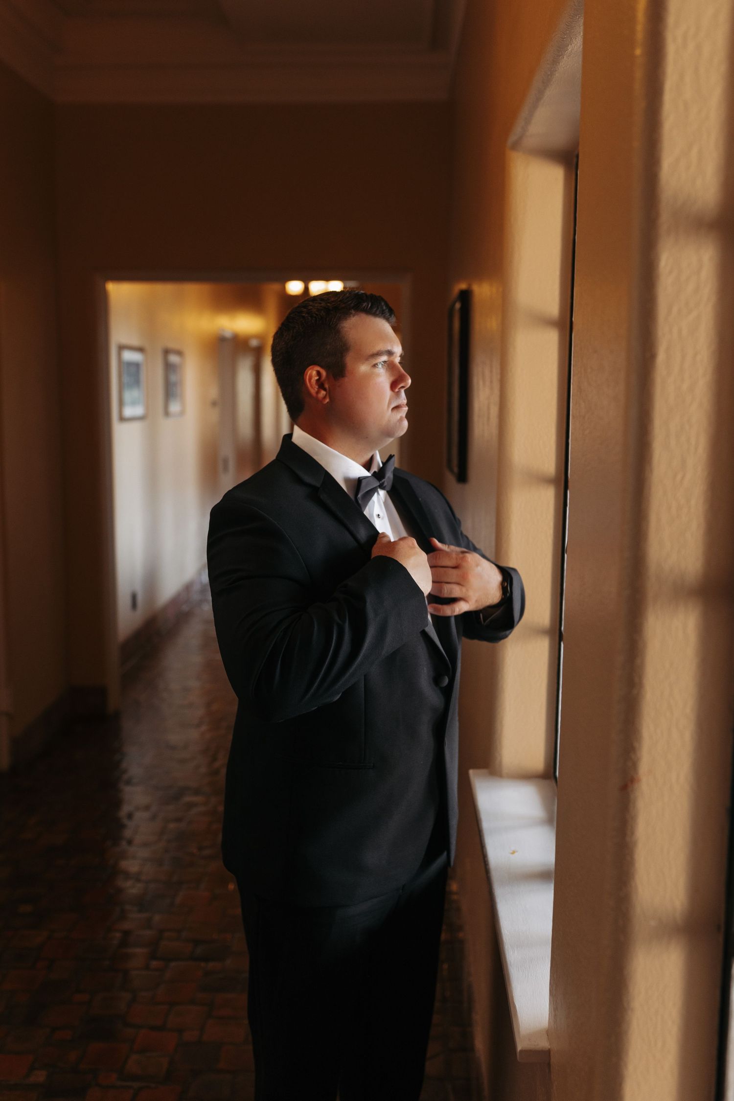 Groom in black tuxedo adjusts bow tie in sunlit hallway with brick floors before wedding.