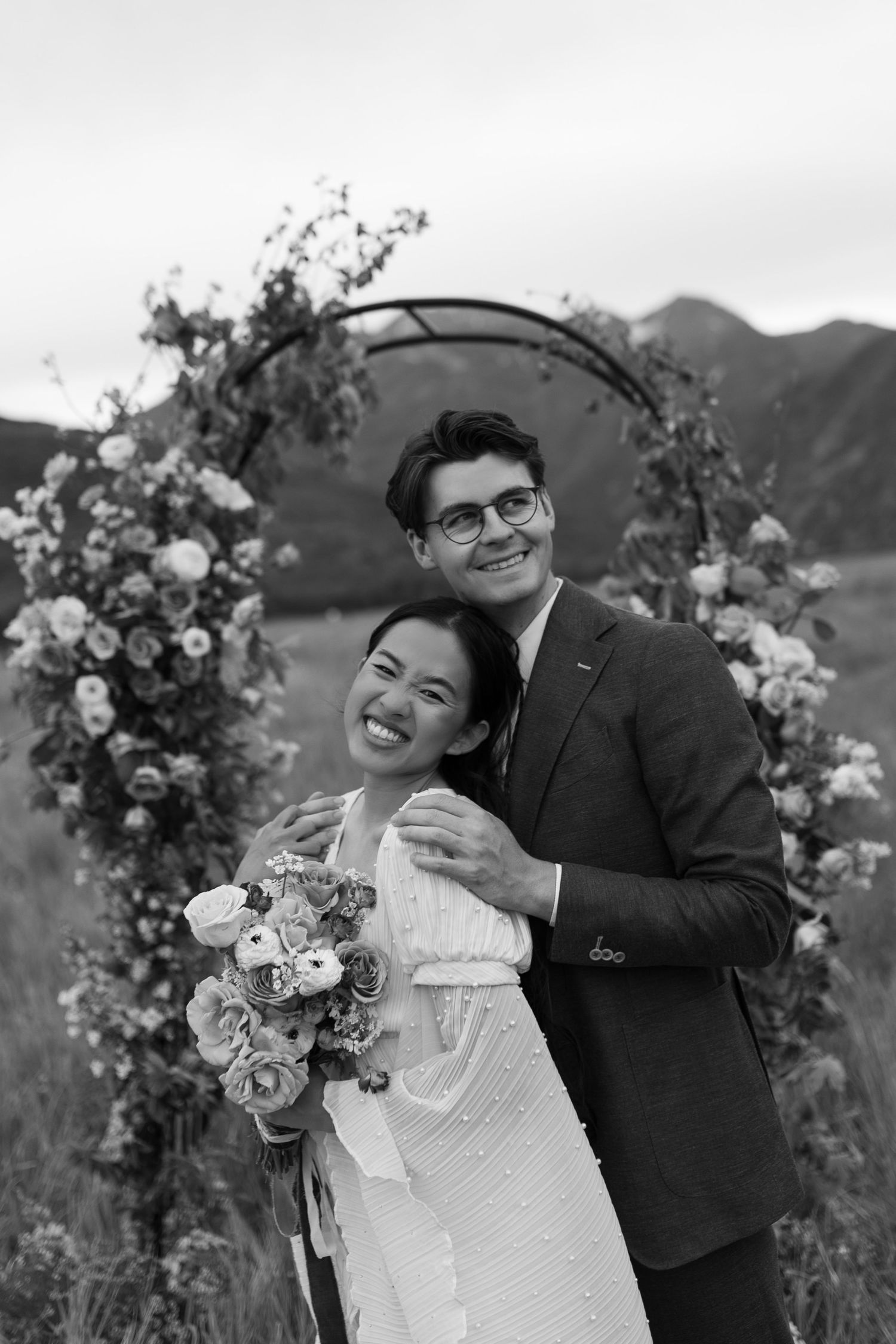 Black and white photo of smiling bride and groom posing before a floral arch with mountain backdrop.
