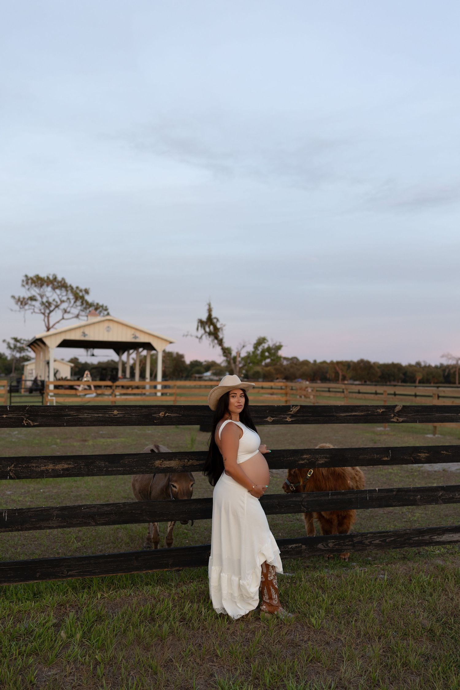 Smiling pregnant woman in white dress and cowboy hat poses by farm fence with cattle.