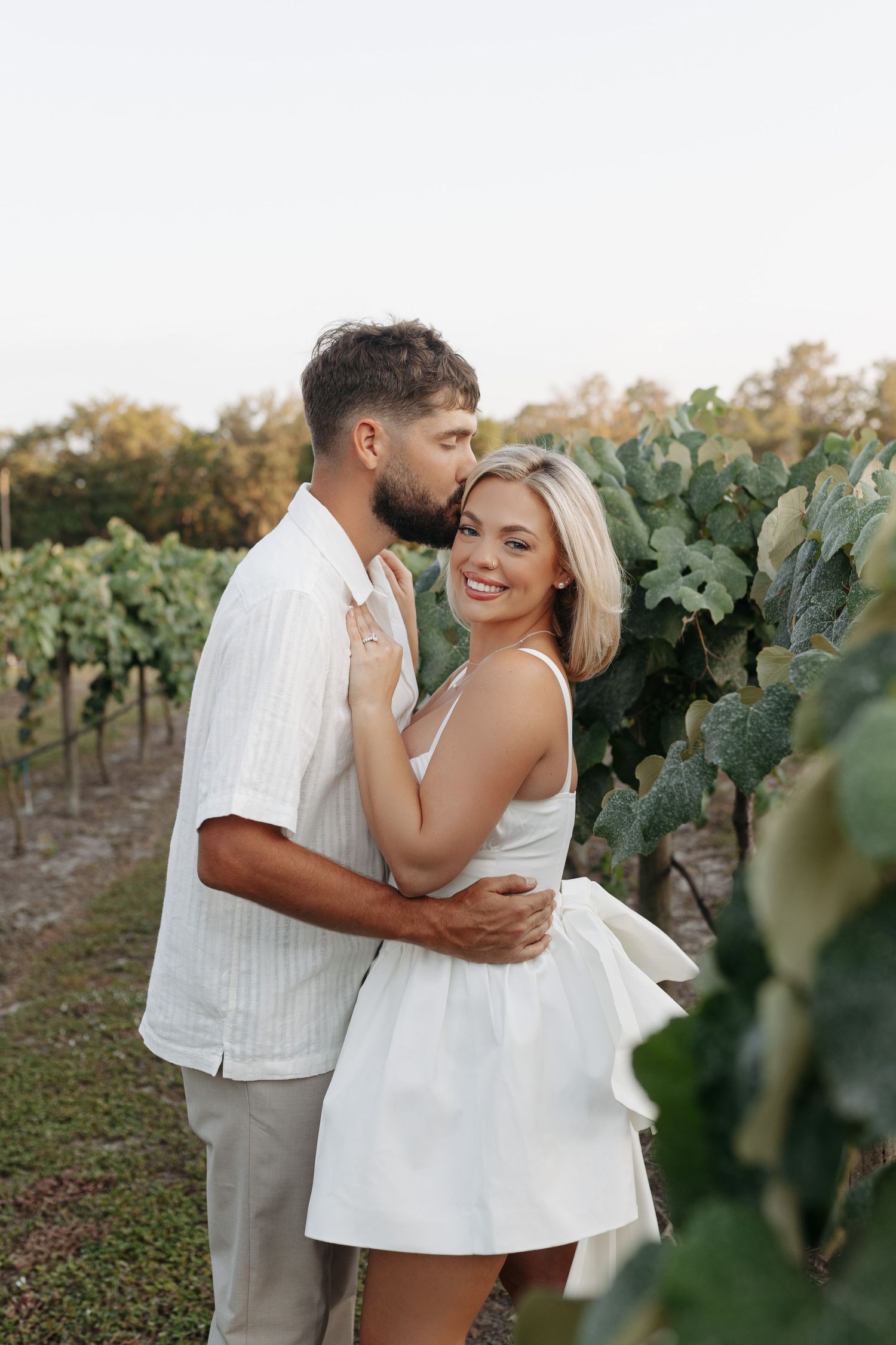 Man kissing woman on cheek while embracing in a lush vineyard.