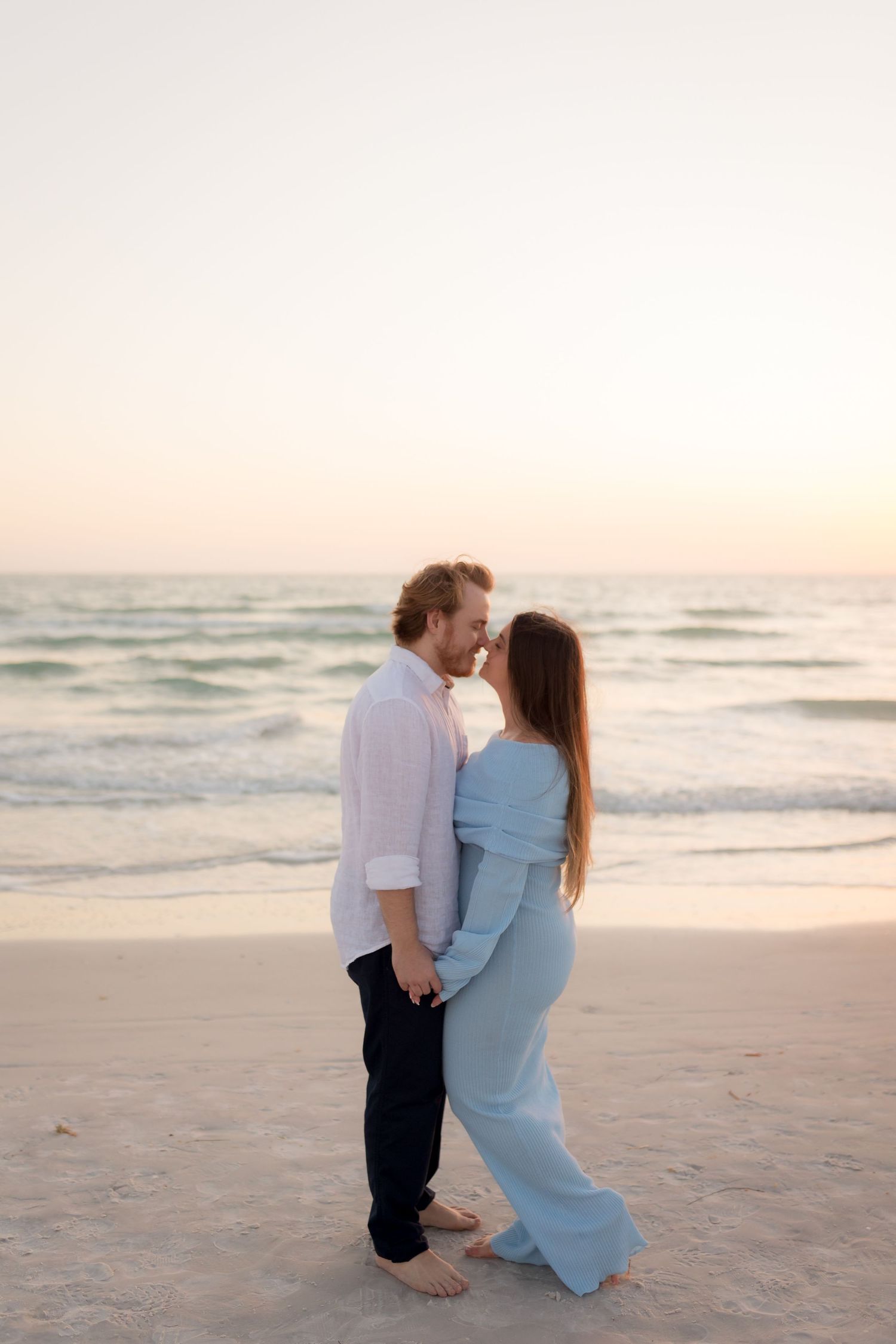 Romantic couple holding hands facing each other on serene beach at pastel sunset in matching blue outfits.