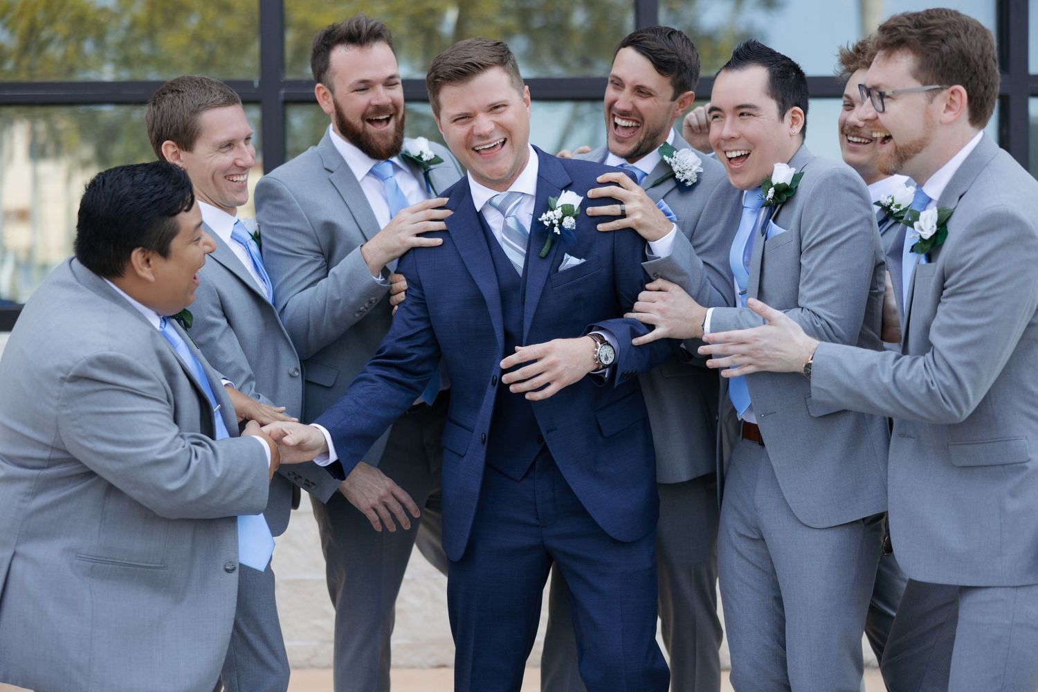 Groom in navy suit laughing with groomsmen in grey suits during fun outdoor wedding party photo.