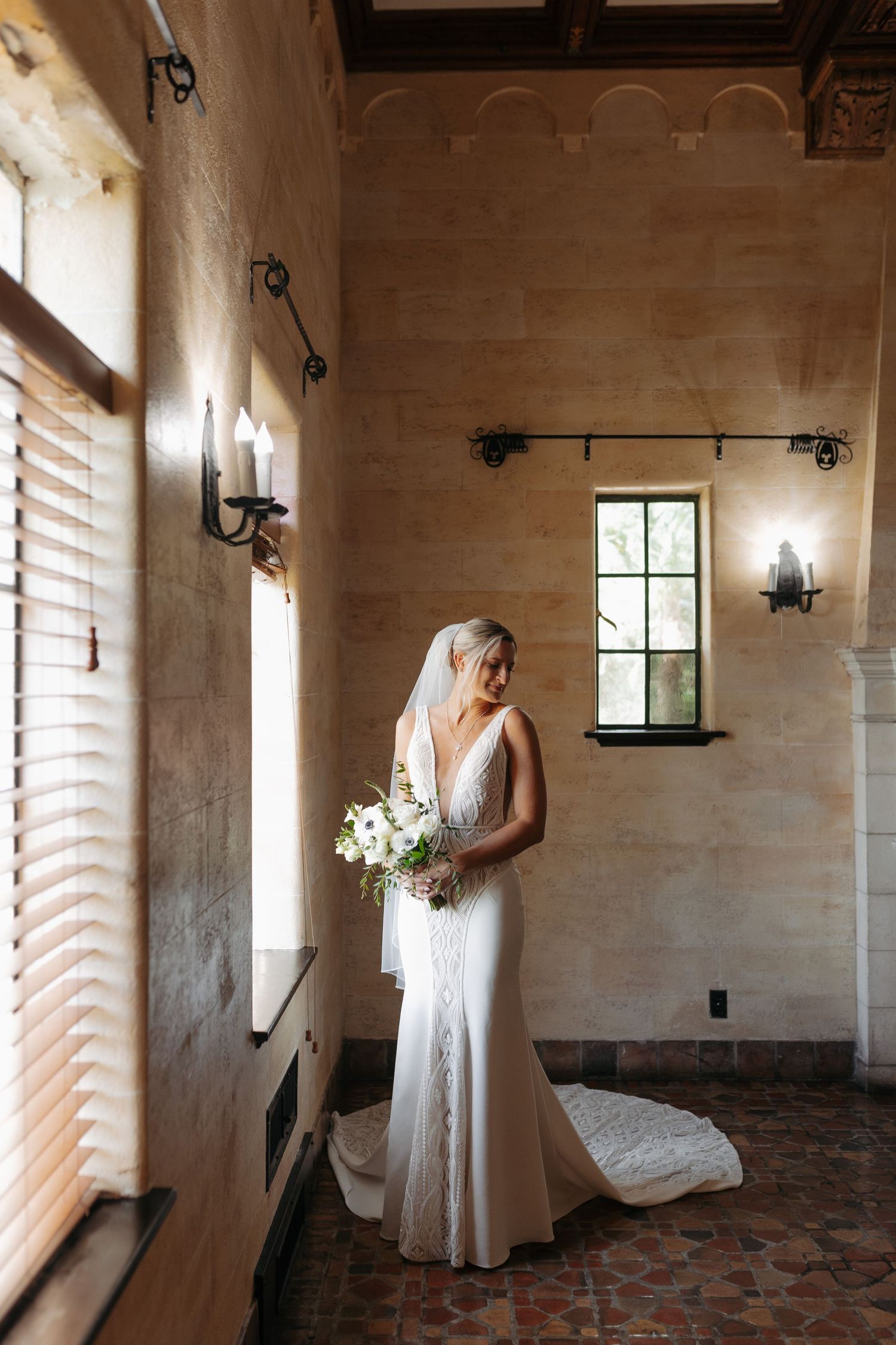 Bride in lace gown holds white floral bouquet while standing in sunlit Spanish-style venue interior.