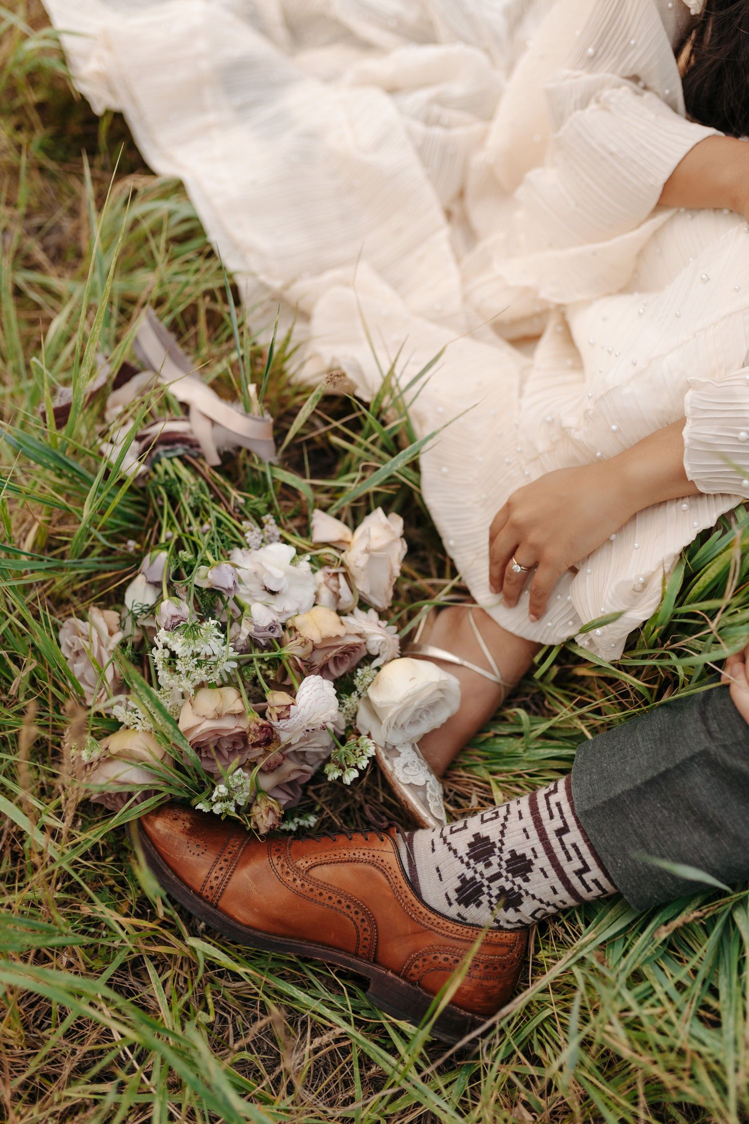 Close-up of bride in white dress sitting in grass with bouquet resting in grooms brown leather brogue shoe.