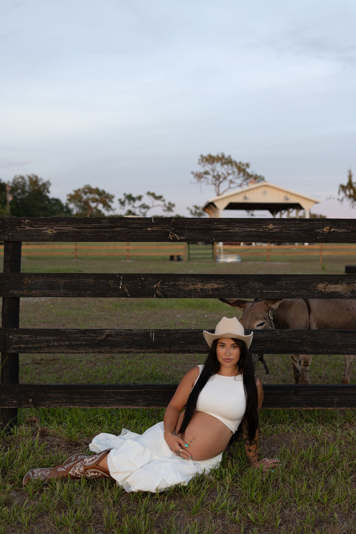 Pregnant woman in white crop top, skirt, cowboy hat, and boots sits against rustic fence on a farm with cattle.