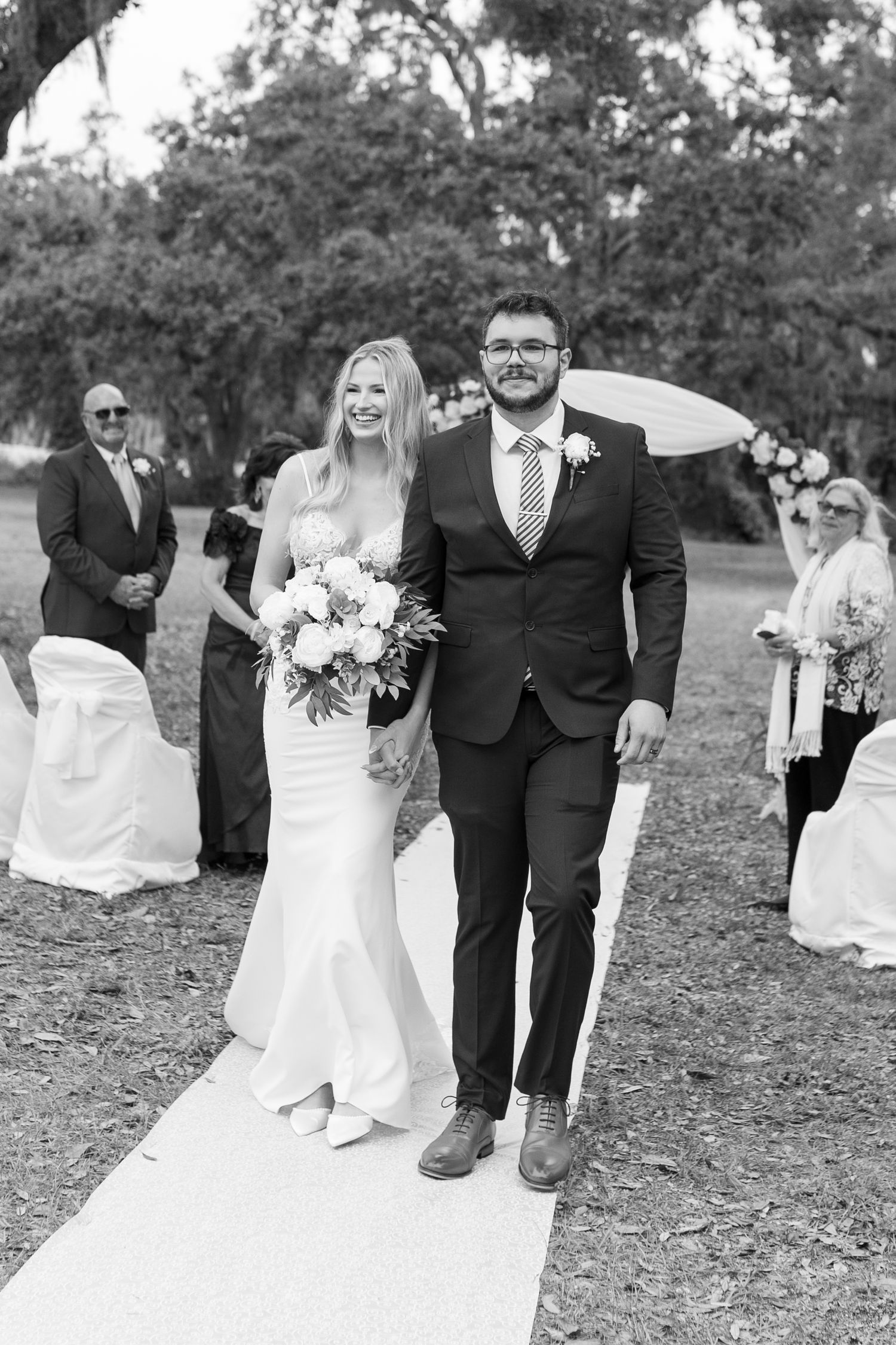 Smiling newlyweds walking down outdoor wedding aisle in classic black and white photography under oak trees.