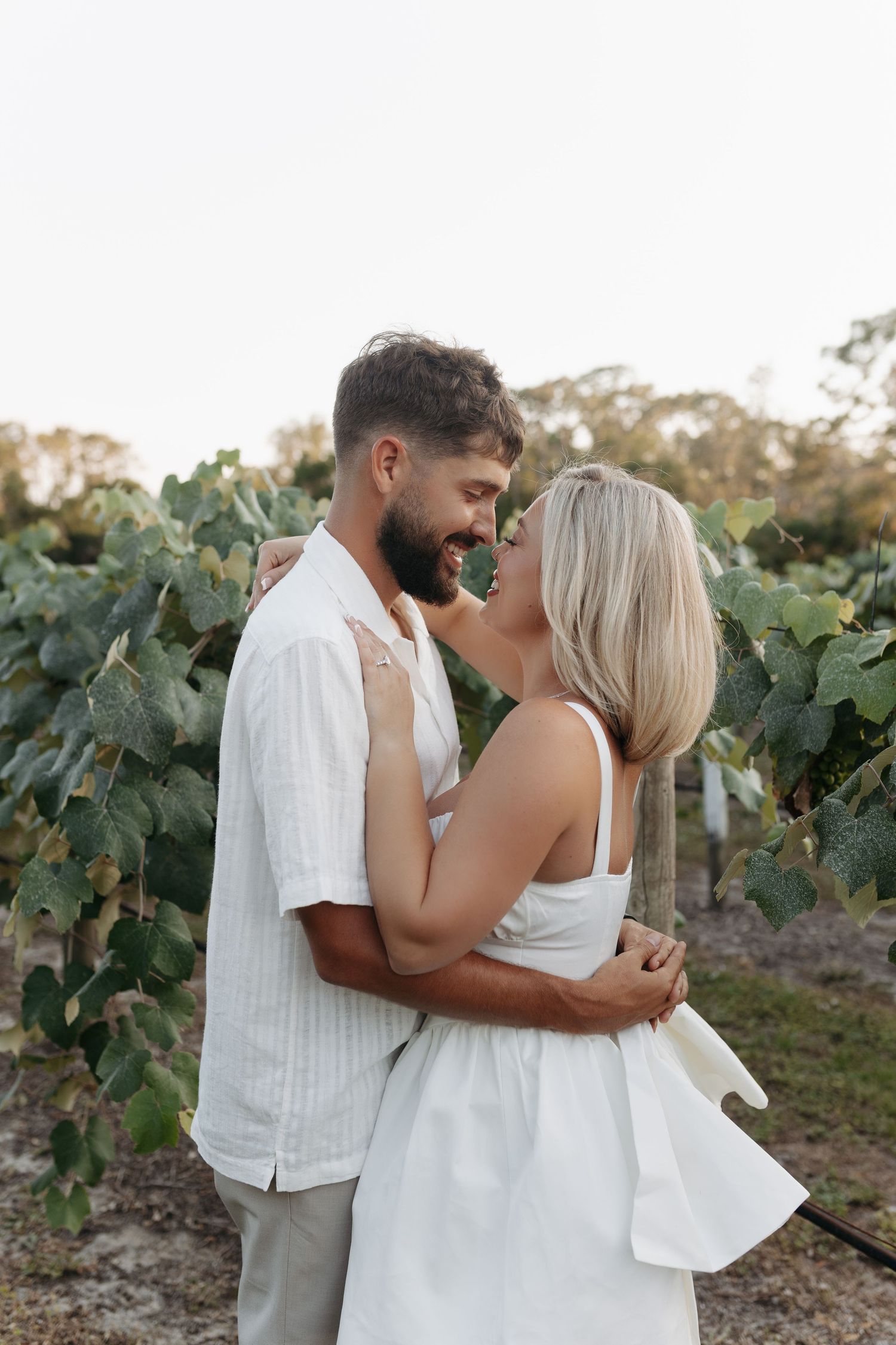 Couple embracing and smiling near green vineyard vines outdoors.