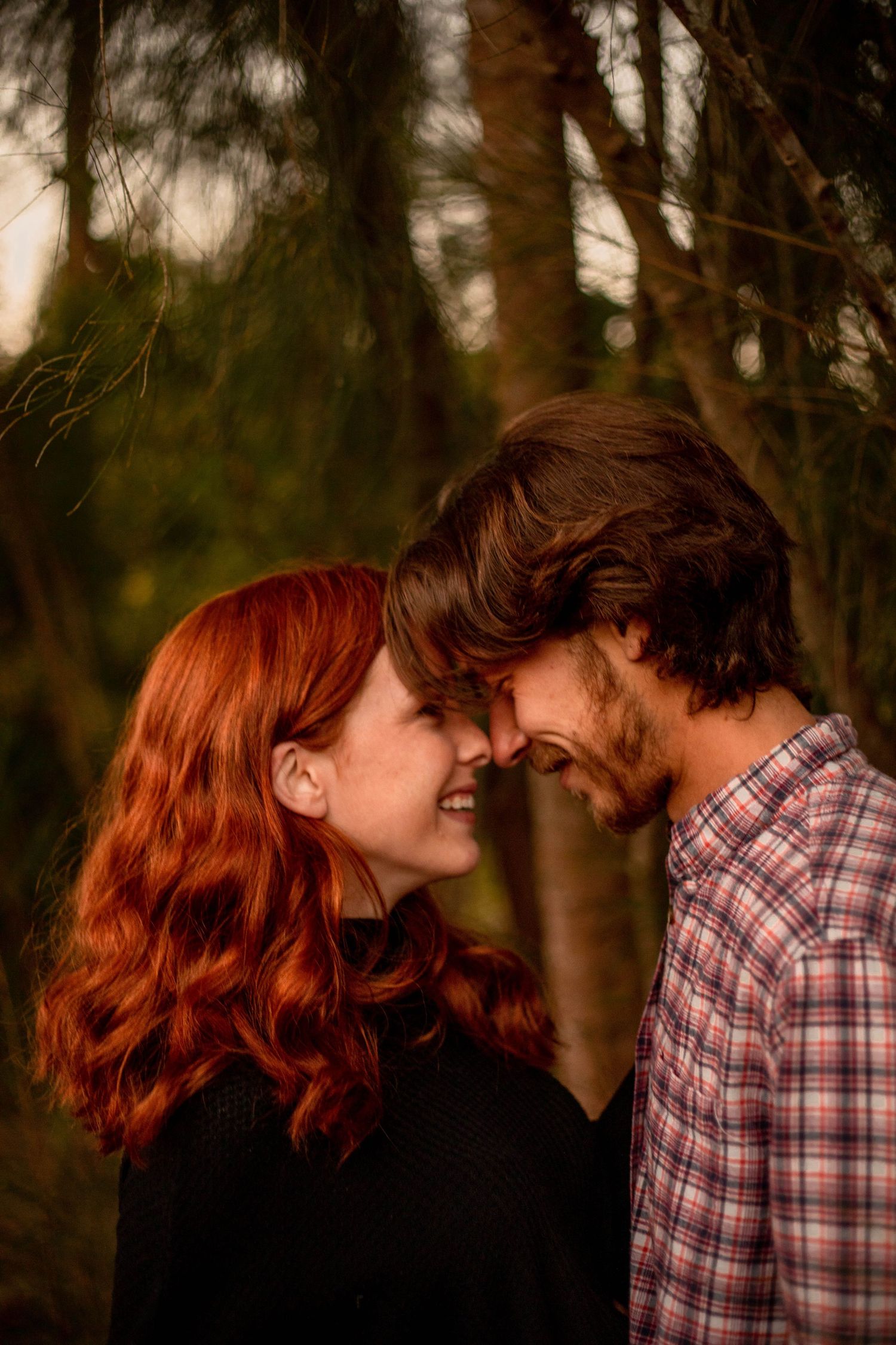 Redhead woman and bearded man touching foreheads and smiling in a romantic forest setting at dusk.