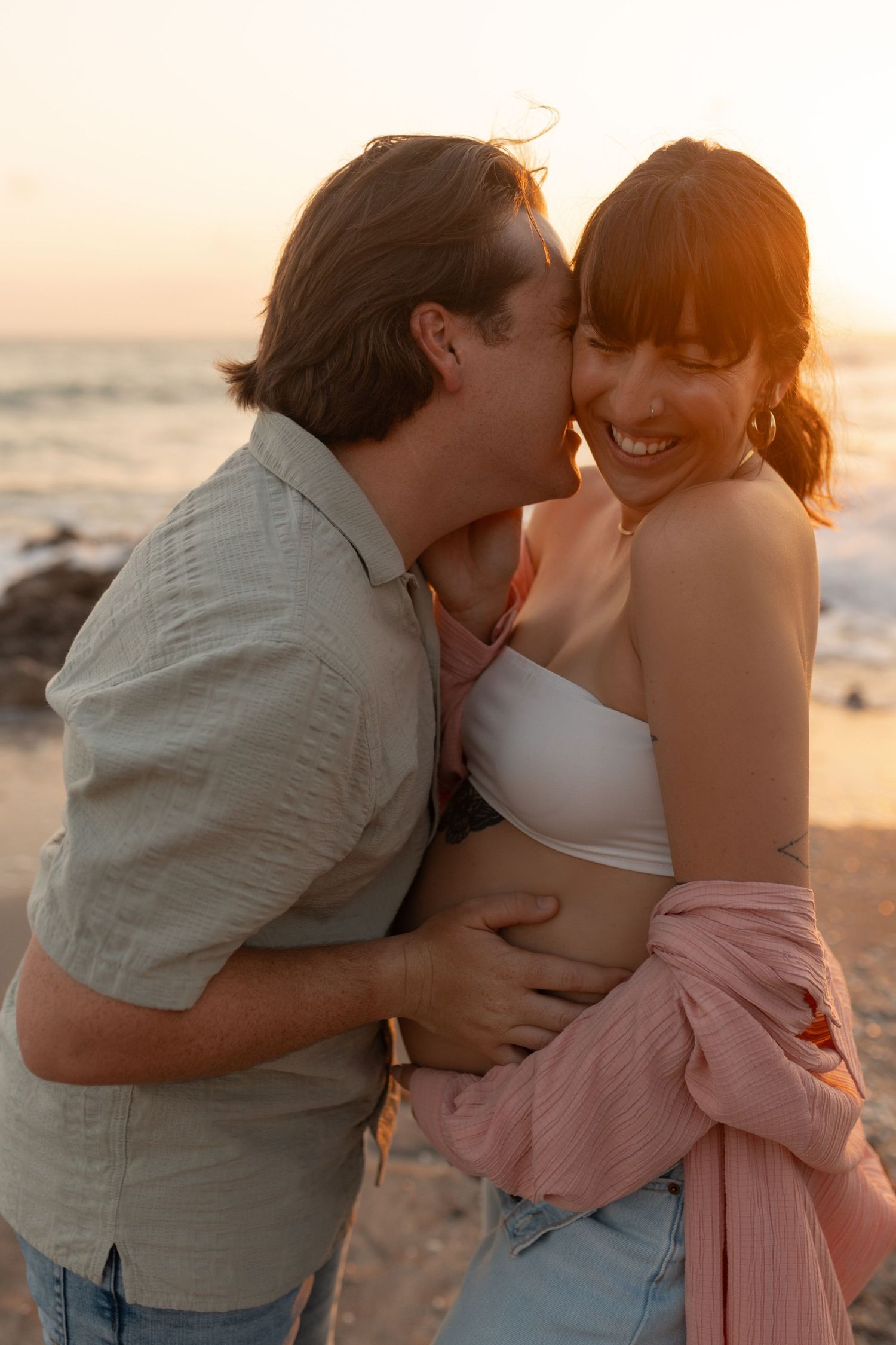 Man kisses pregnant woman's cheek as she laughs on beach at sunset, both sharing a tender embrace.
