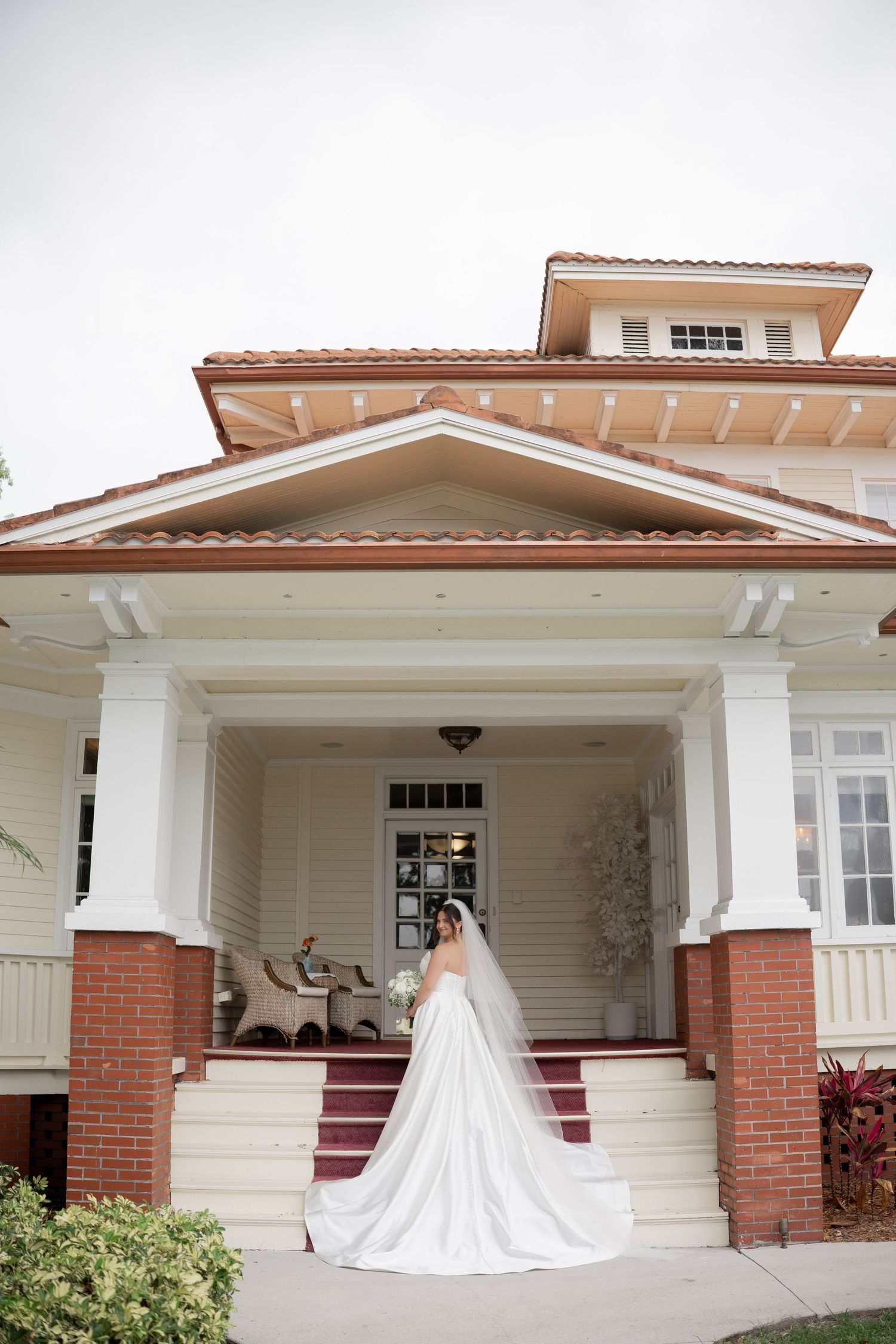Bride in long white gown and veil poses on steps of elegant white and brick wedding venue.