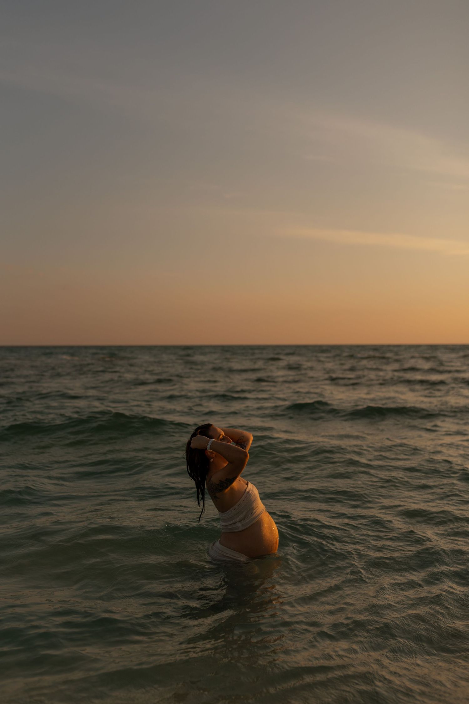 A woman stands waist-deep in ocean waves at sunset, hands raised to her dark wet hair.
