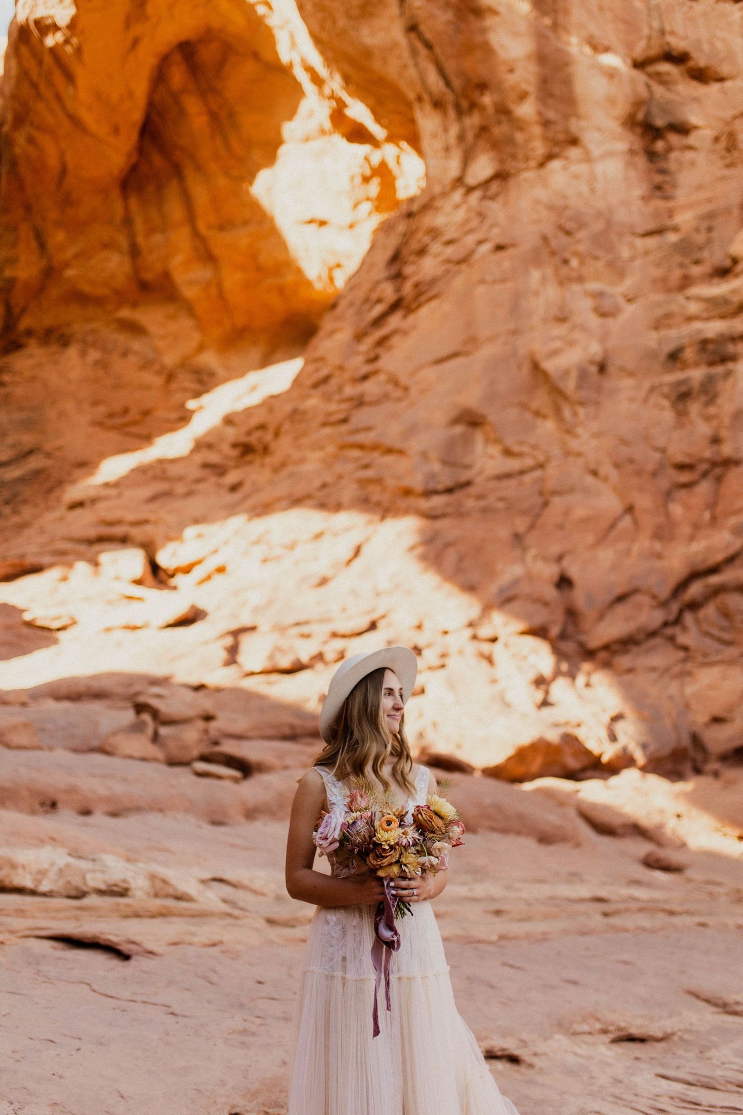 Elegant bride in flowing gown and hat holding bouquet beside a stunning red sandstone arch formation.