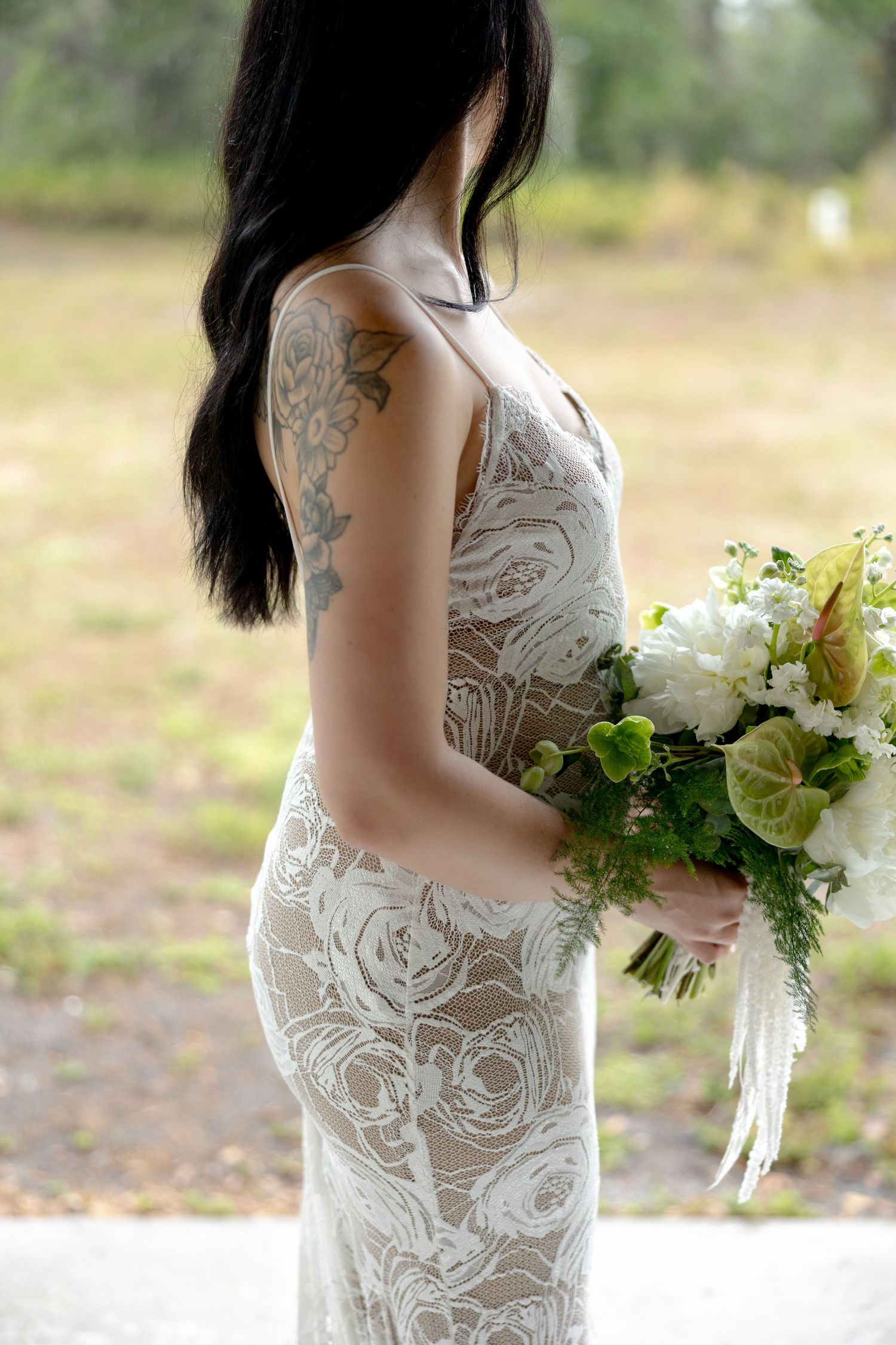 Side profile of tattooed bride in fitted lace wedding dress holding elegant white floral bouquet.