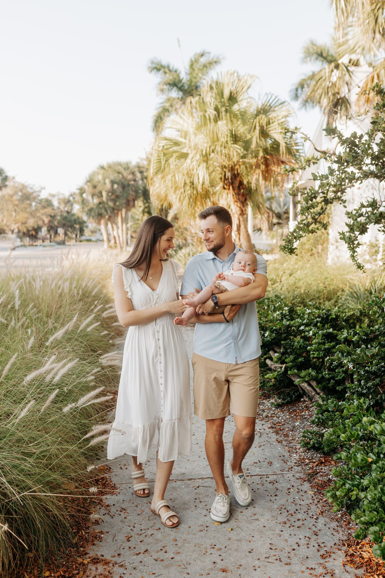 Young family strolling along a sunny palm-lined path, father holding newborn baby in Florida.