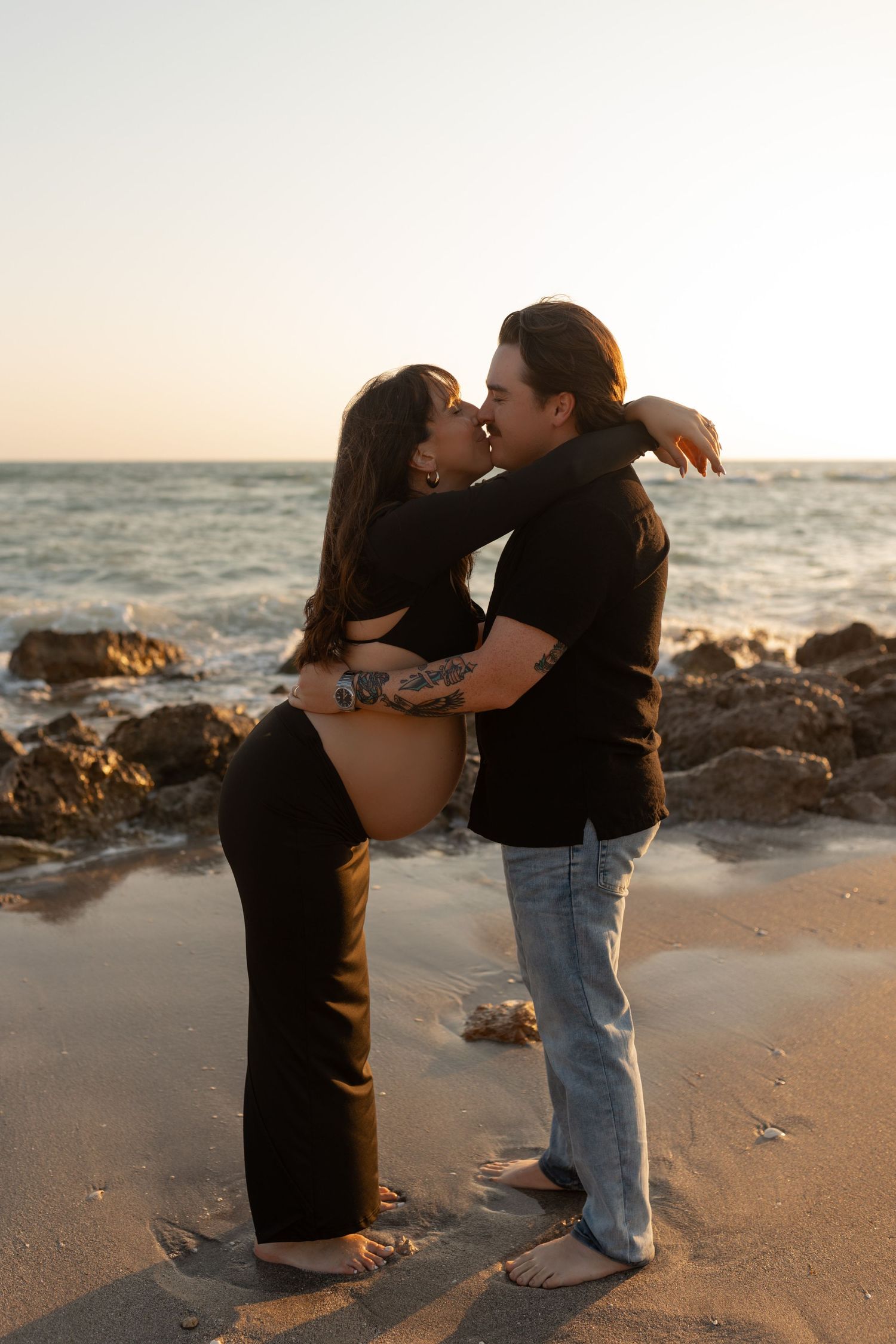 Pregnant couple shares a kiss on rocky beach at sunset with ocean waves behind them.