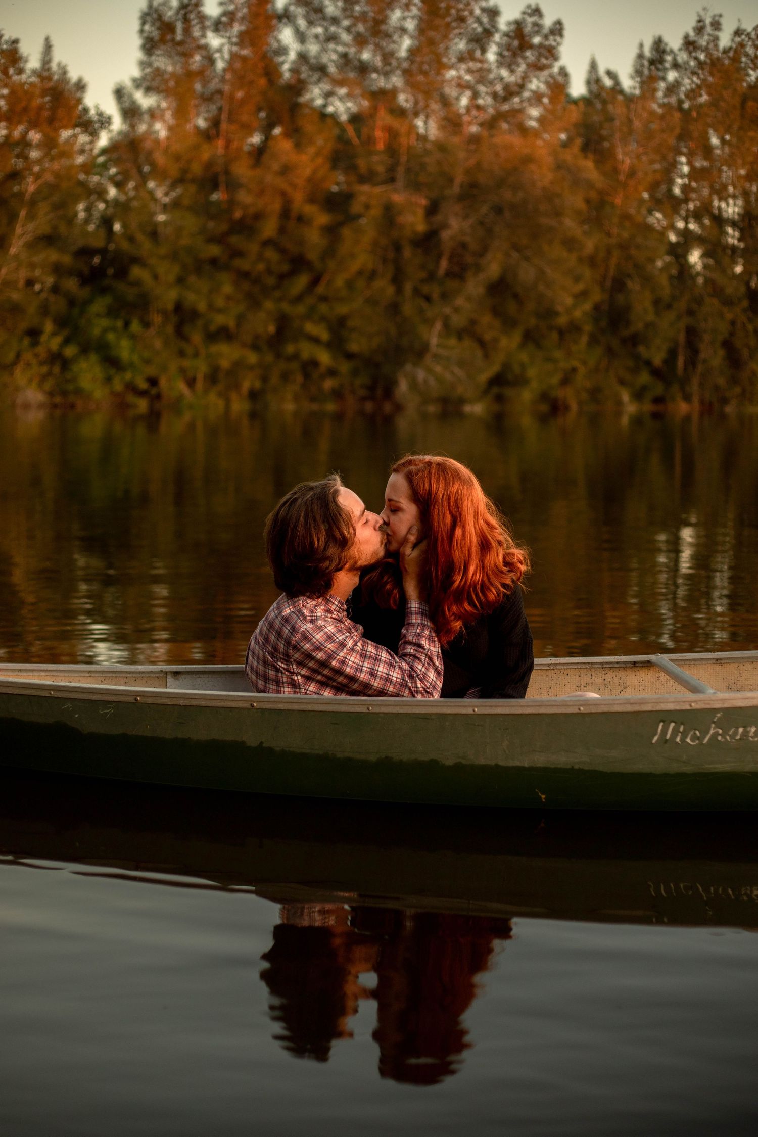 Couple sharing a kiss in a canoe on a calm lake surrounded by autumn foliage at dusk.