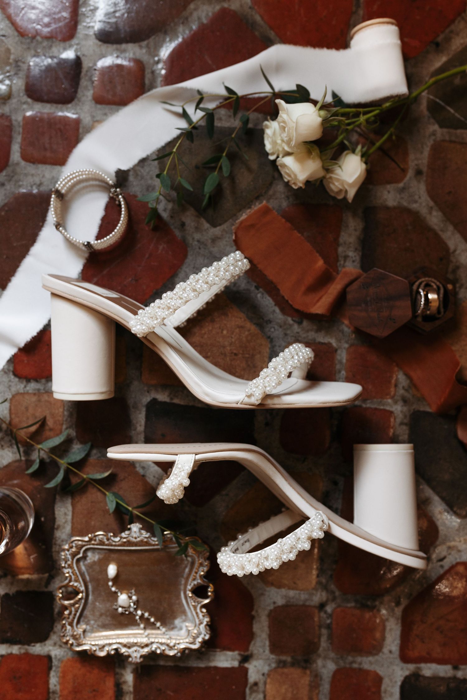 Flat lay of white pearl-strap bridal heels, bracelet, white roses, and accessories on rustic brick surface.
